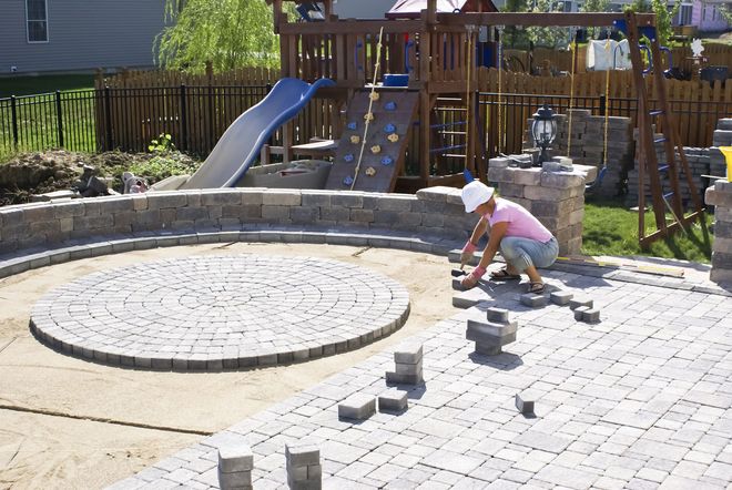 A person in a pink shirt and white hat installs stone pavers in a backyard patio near a playground set.
