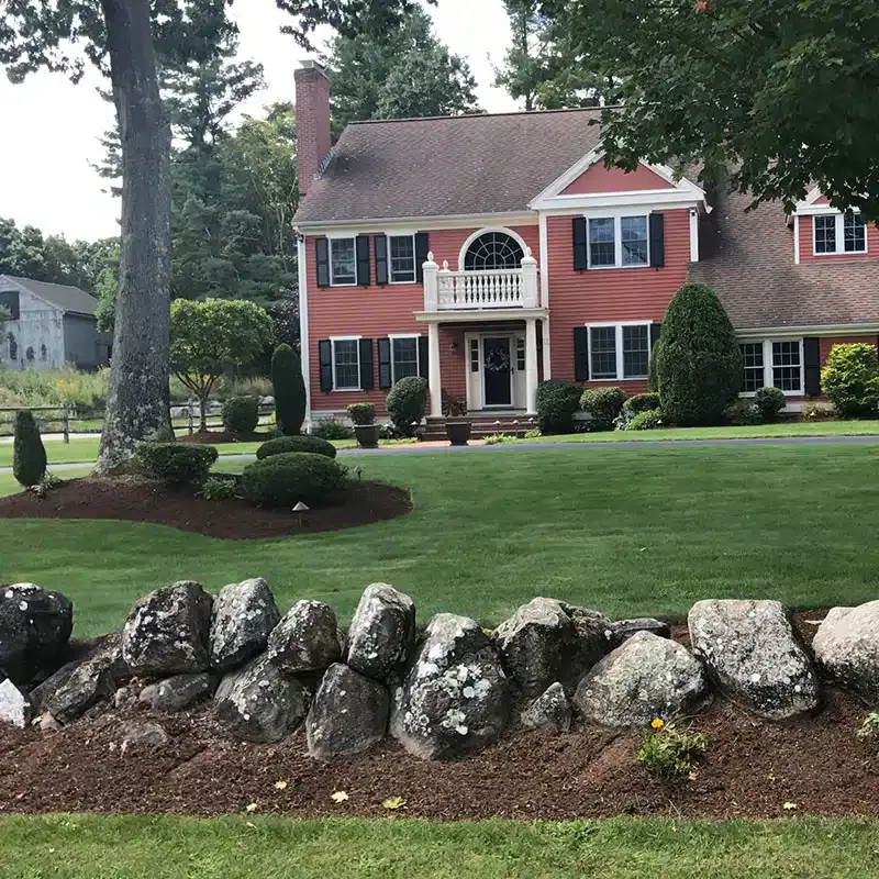 A two-story red colonial house with a stone wall in the foreground and a landscaped green lawn.
