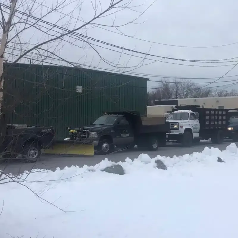 A line of dark-colored utility trucks, some with snowplows attached, parked in front of a green building in the snow.