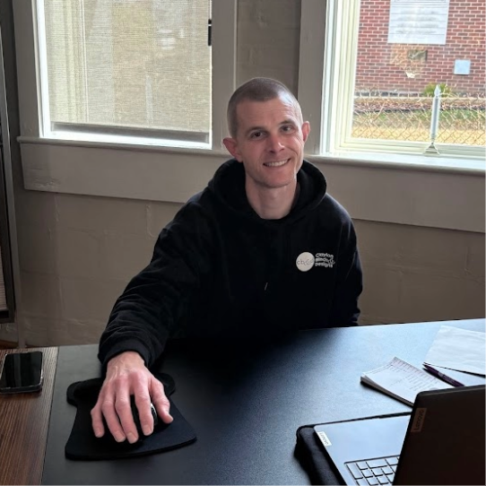 Man in black hoodie smiles at desk with mousepad, laptop, and phone near window.
