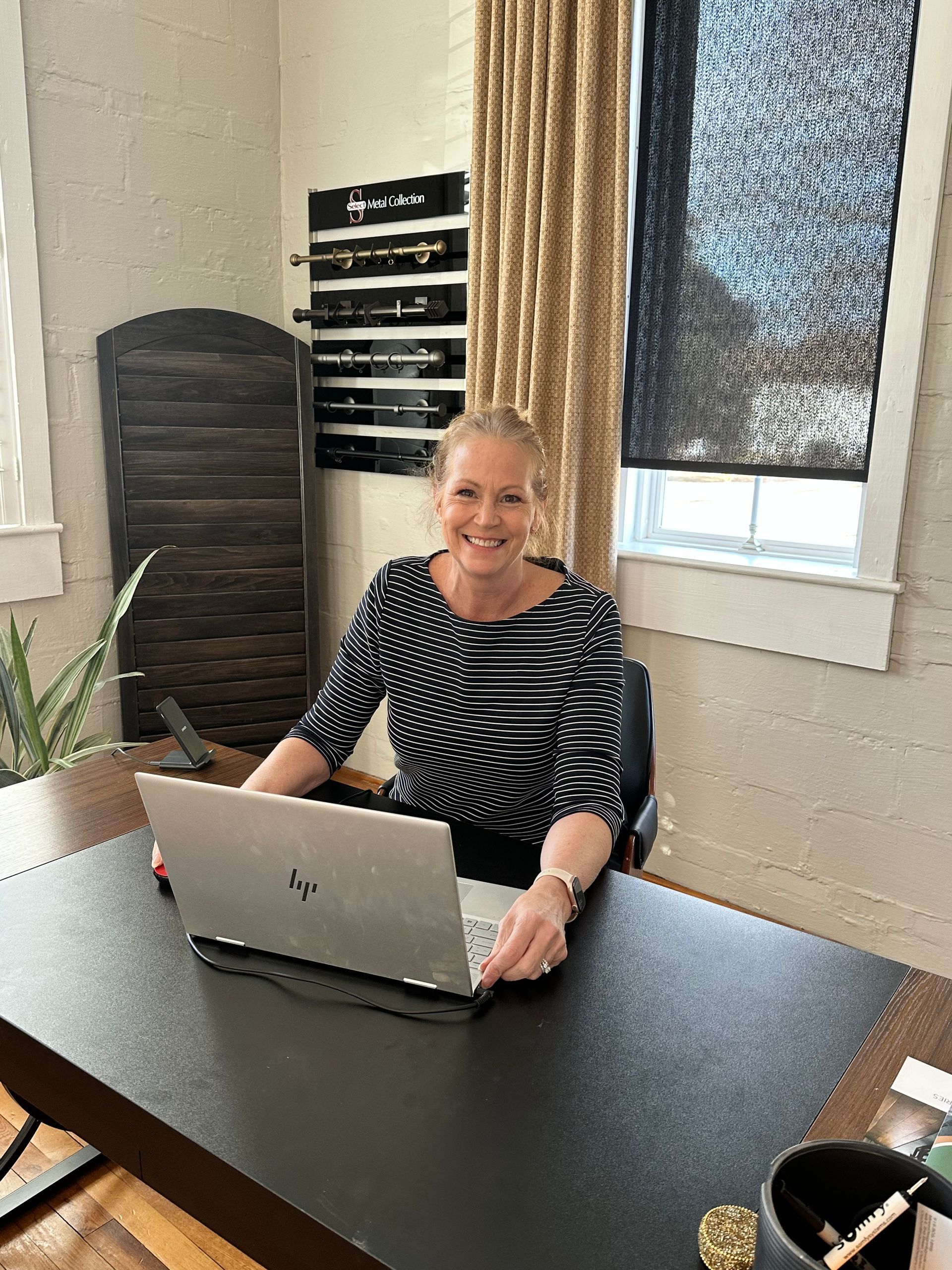 Woman smiles, seated at a desk, using a laptop in an office setting.