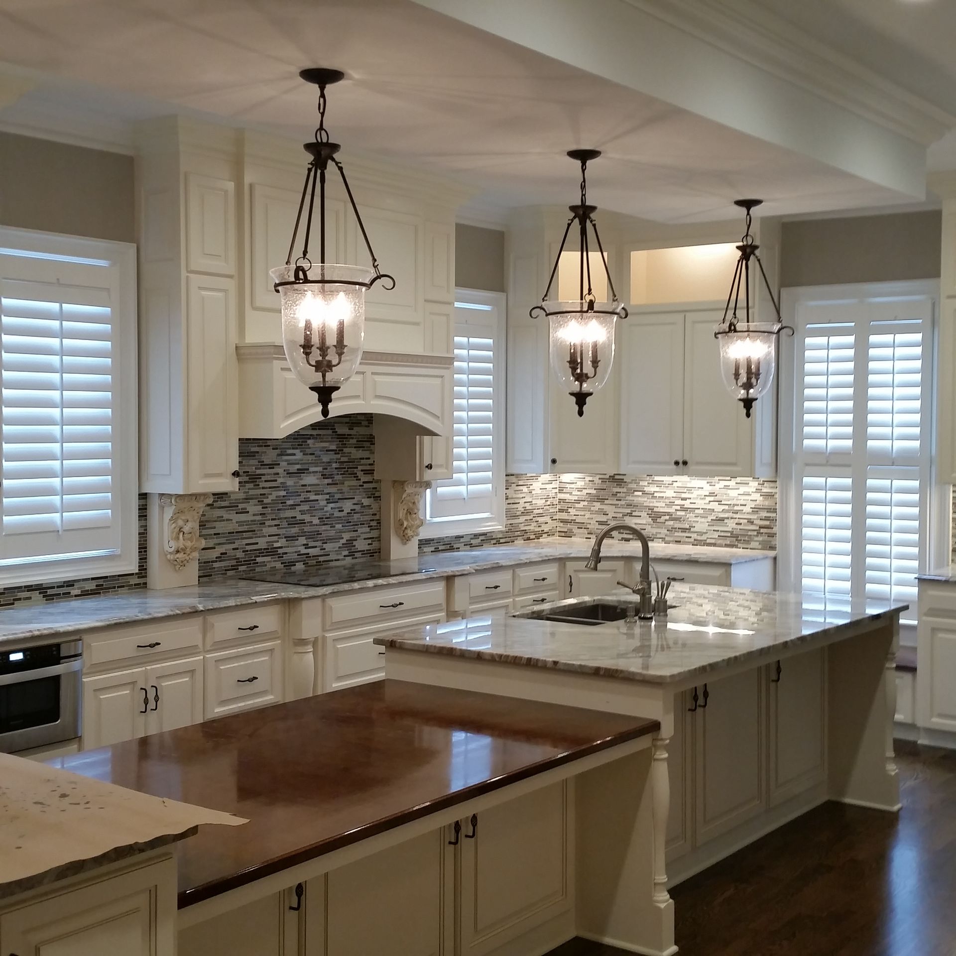 Cream-colored kitchen with island and three hanging lanterns. White cabinets, granite counters, wood floors, and window shutters.