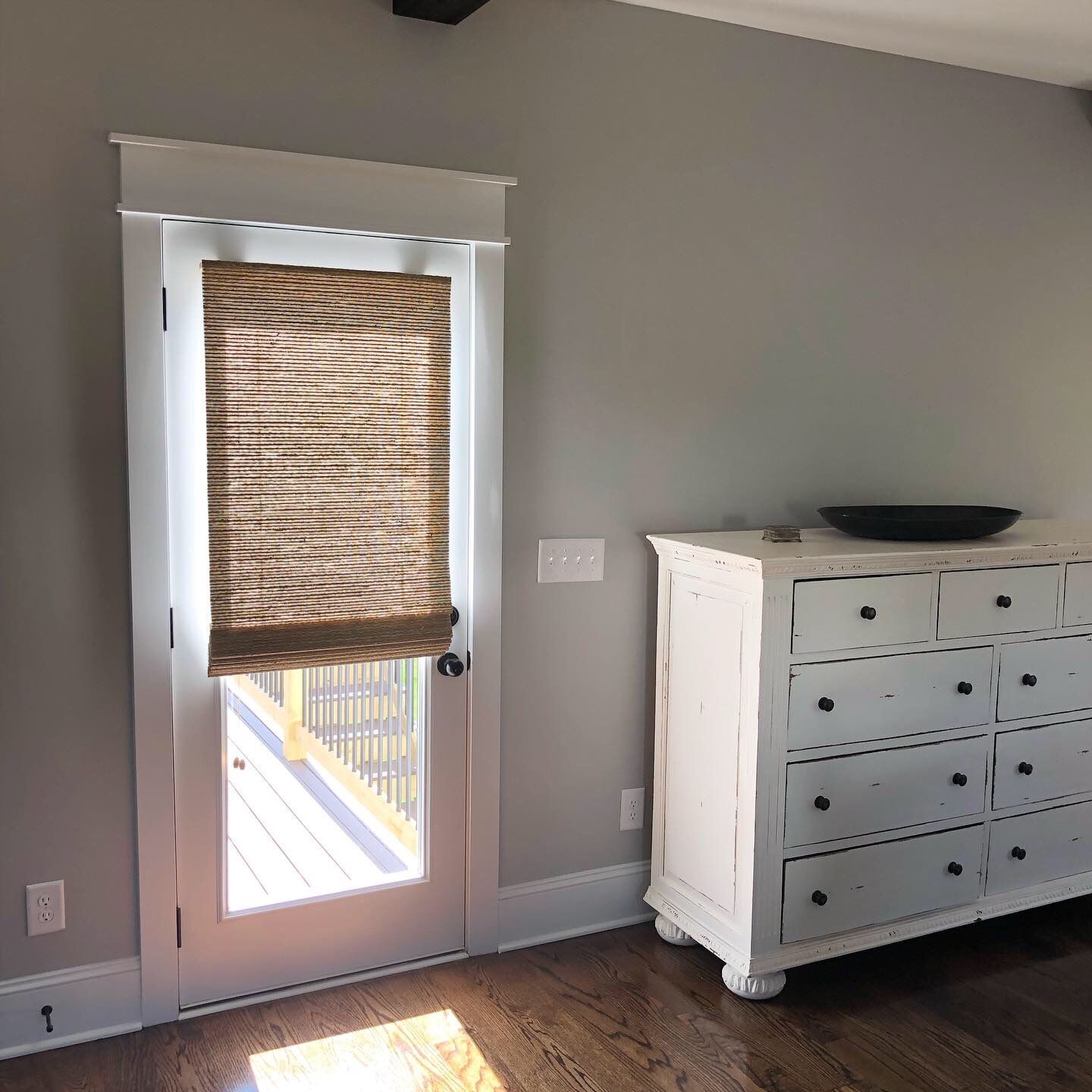 Door with woven shade, gray walls, white dresser, wood floor, and sunlight.