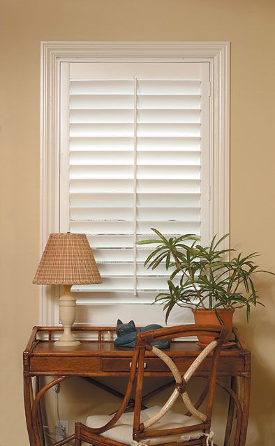Desk with lamp and plant in front of a window with white shutters, in a room with light beige walls.