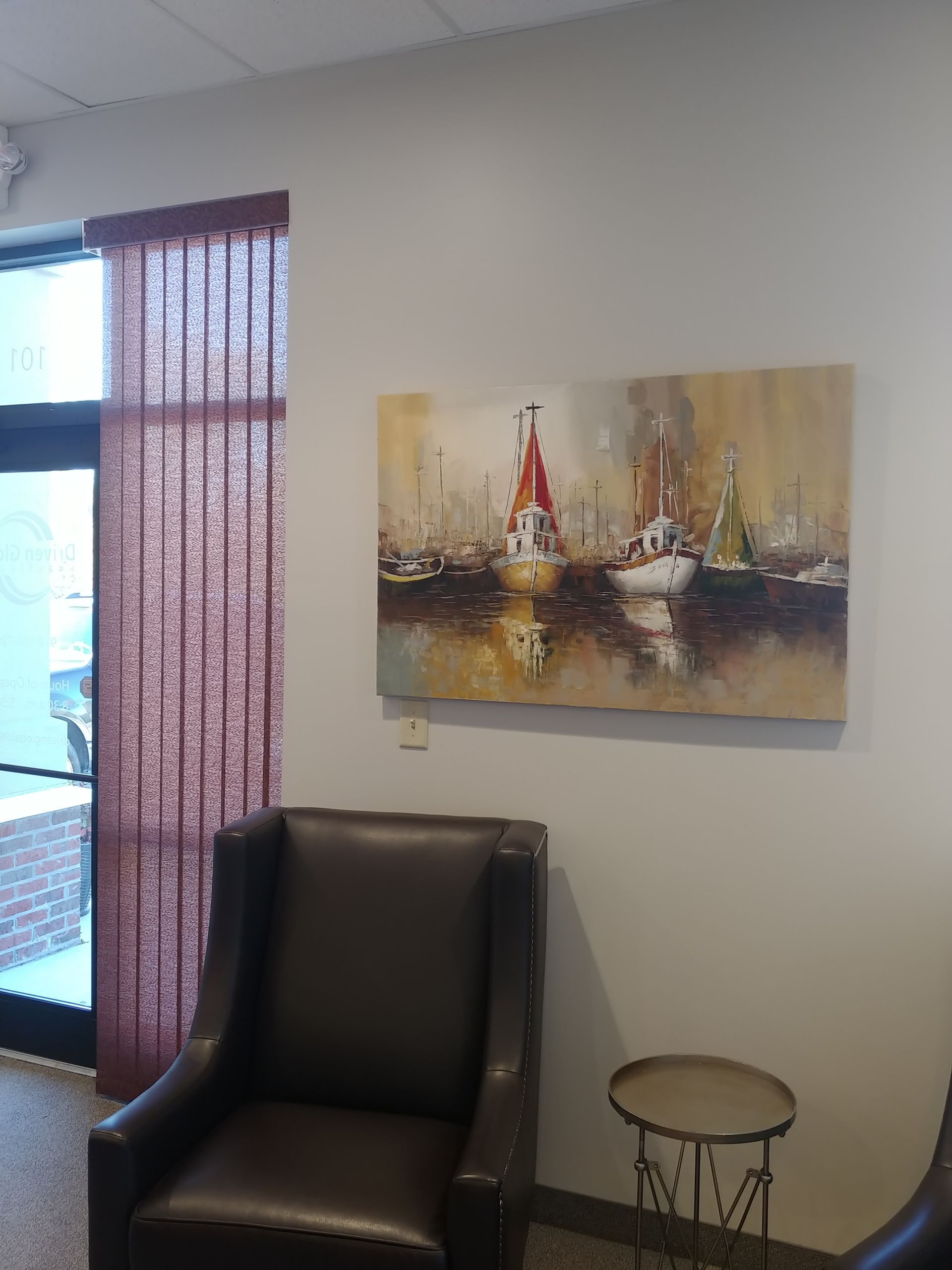 A brown leather armchair, a painting of boats, and a small table in a room with a window and maroon vertical blinds.