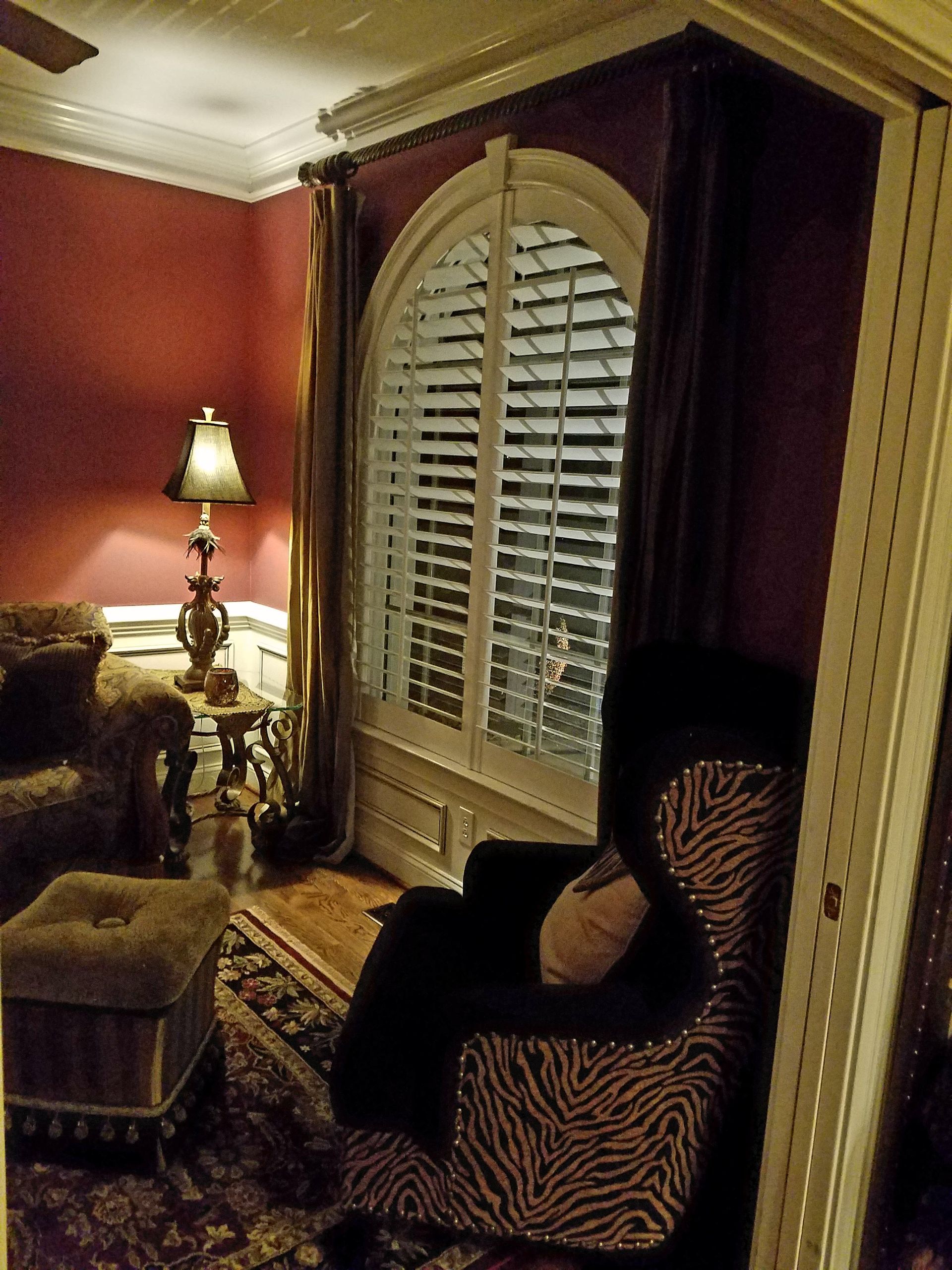 Cozy room with red walls, patterned armchair, window with shutters, and a lamp on a side table.