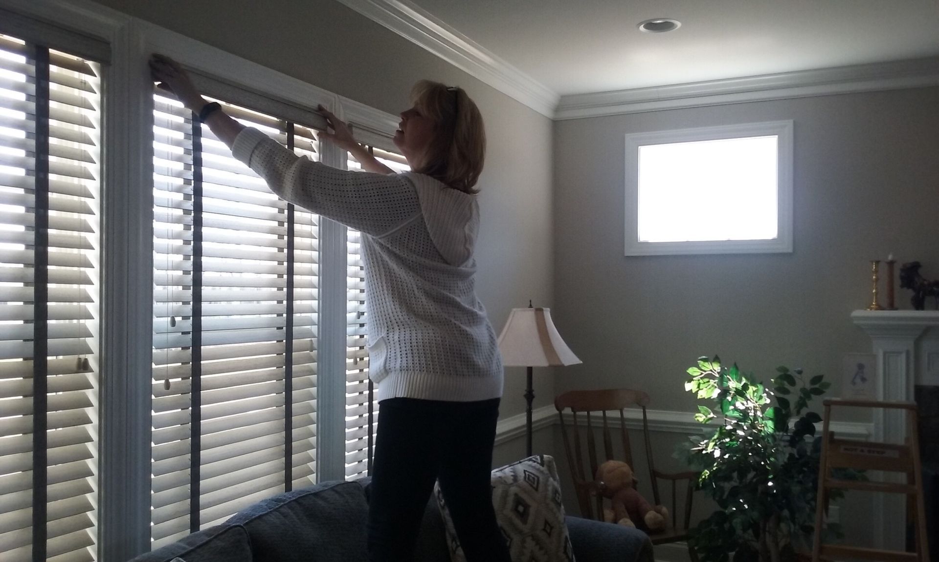 Woman adjusting blinds in a sunlit room. She is standing on a sofa.