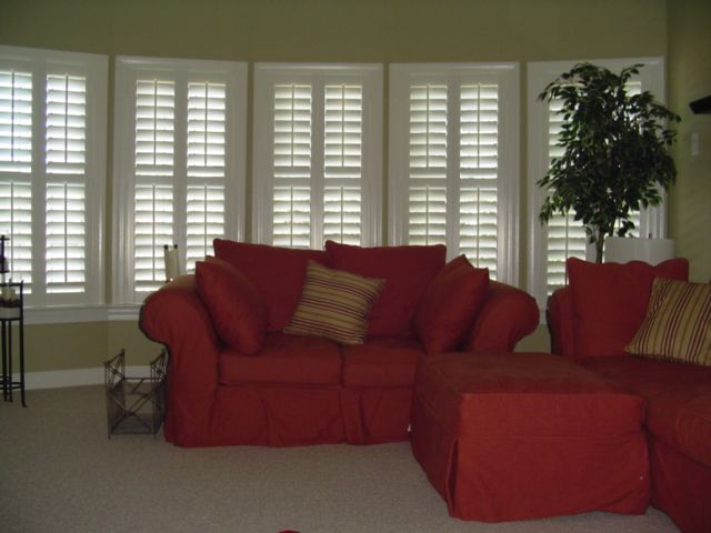 Red sofa and ottoman in front of shuttered windows; artificial plant in the corner.