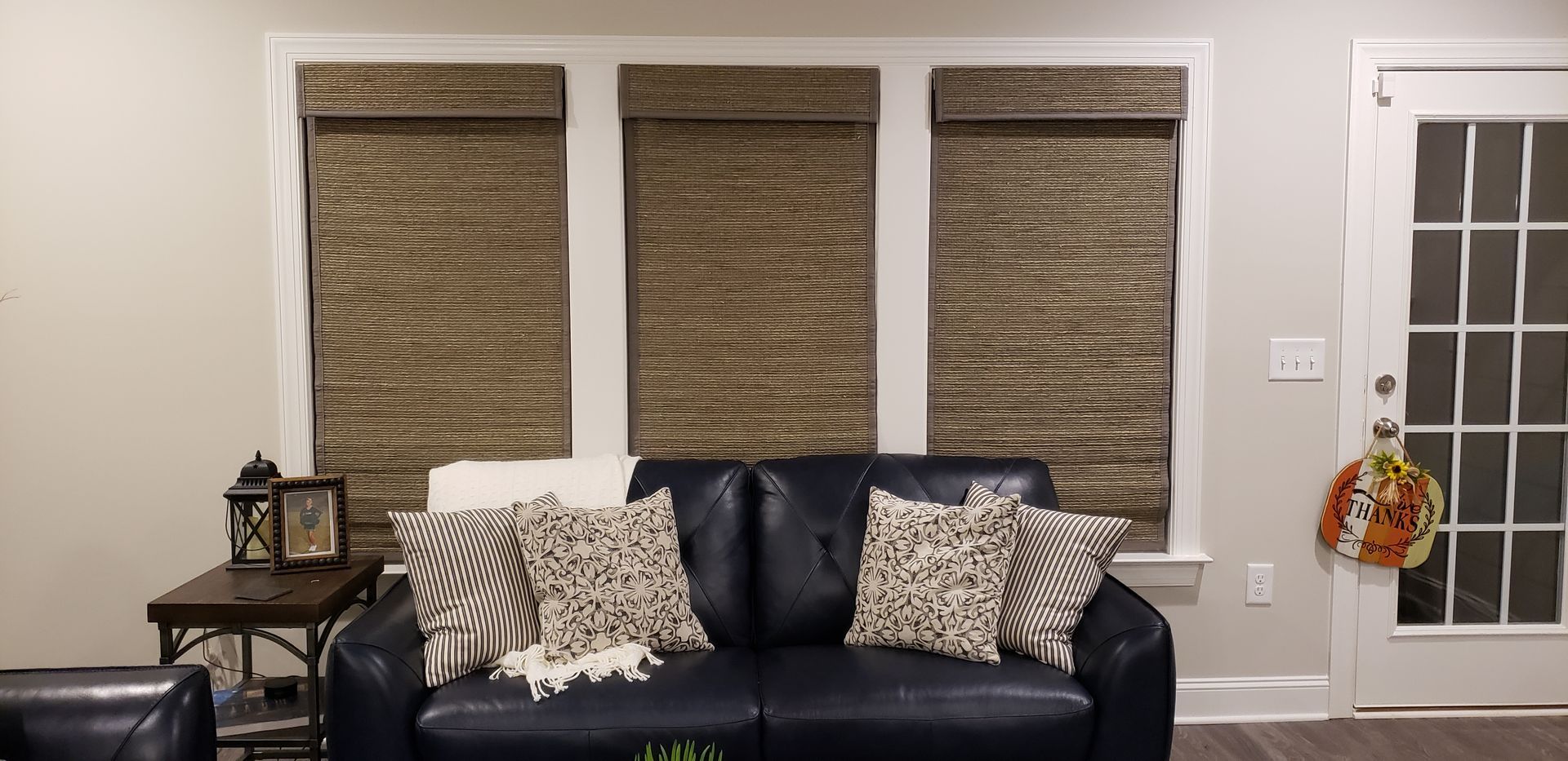 Living room with three windows with woven blinds, a black leather couch, and a glass door with a fall wreath.
