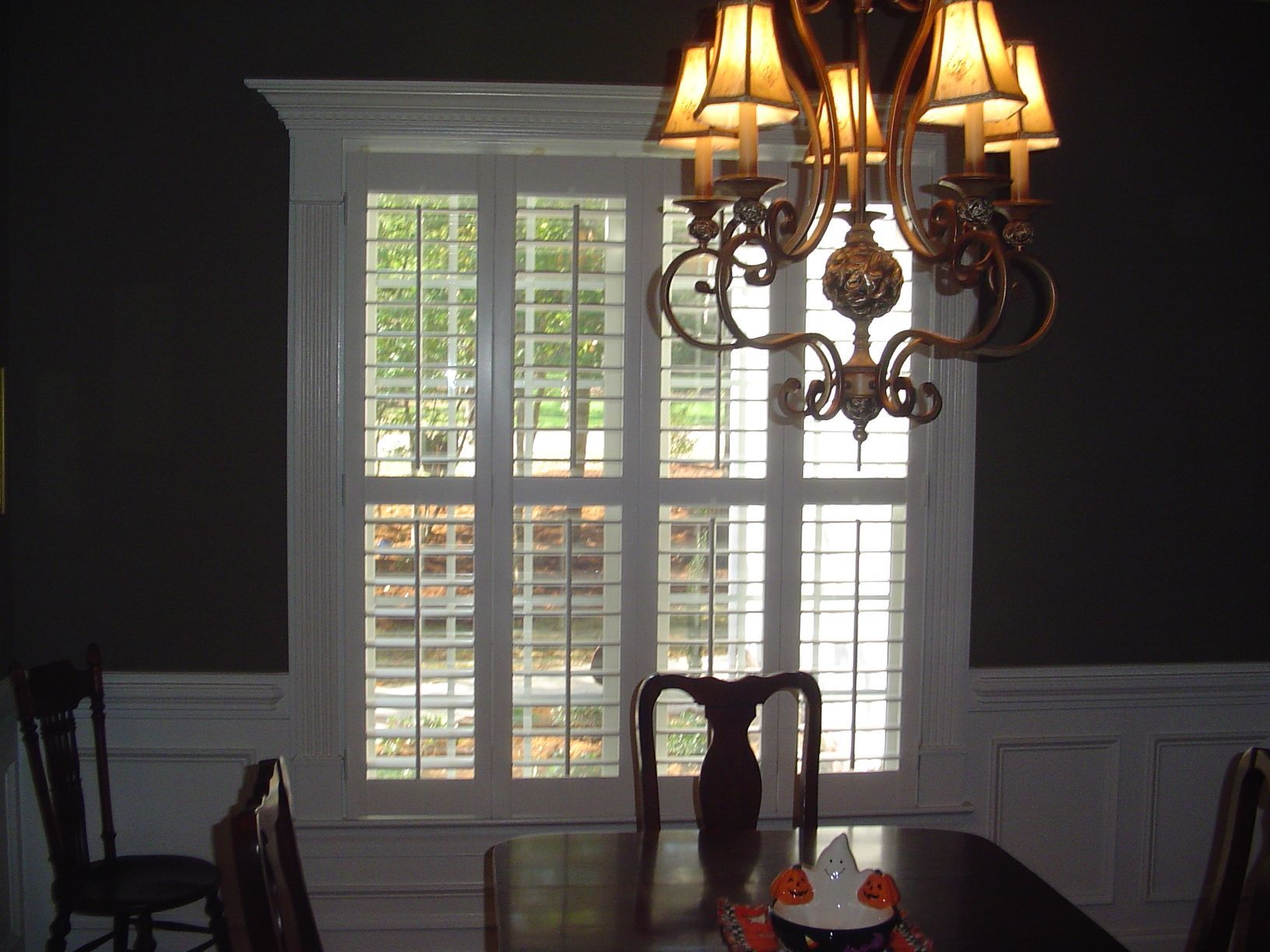 Dining room with white shuttered window, chandelier, dark walls, and table with chairs.
