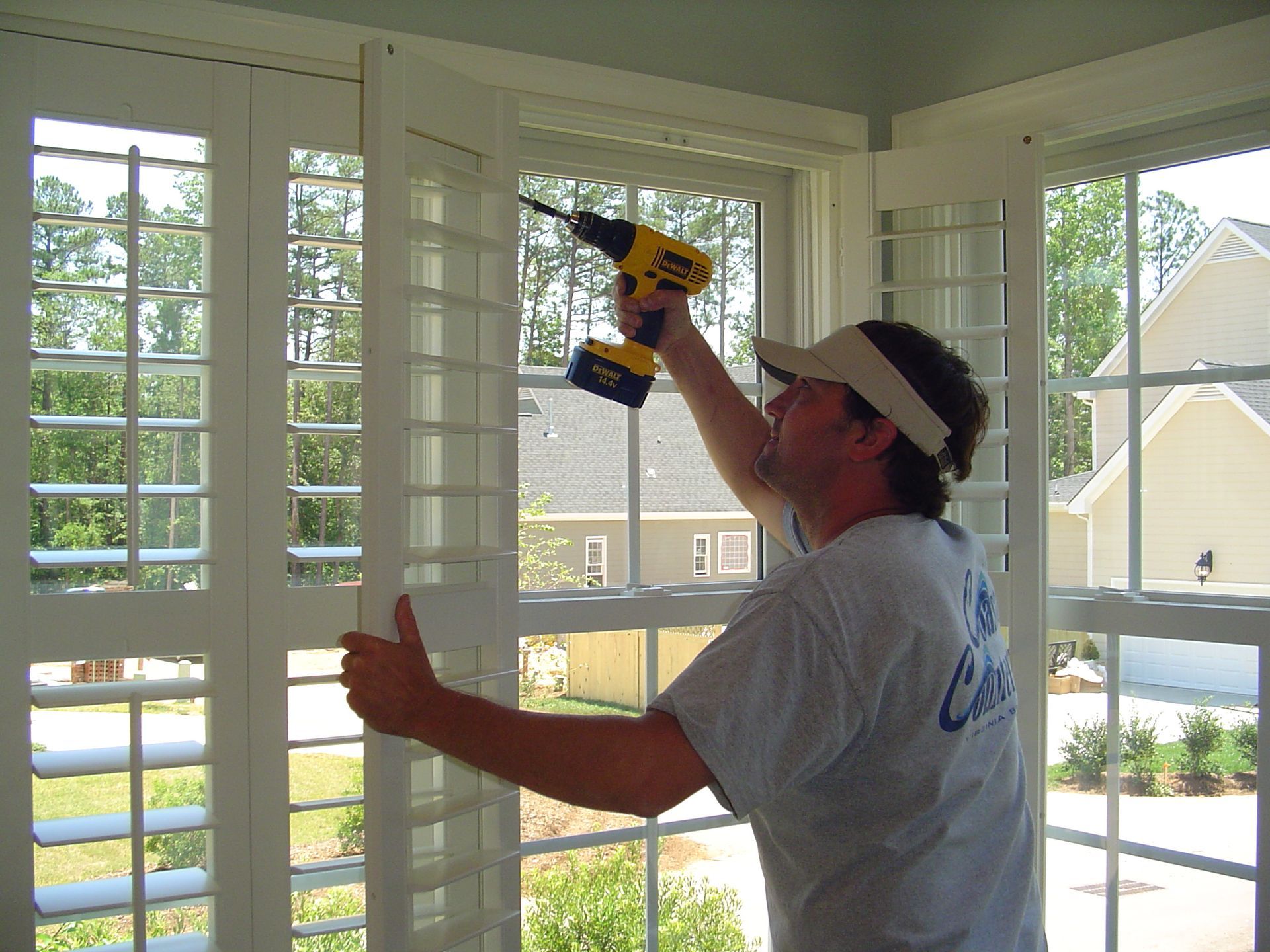 Man installing white shutters with a drill in a windowed room; bright sunlight.