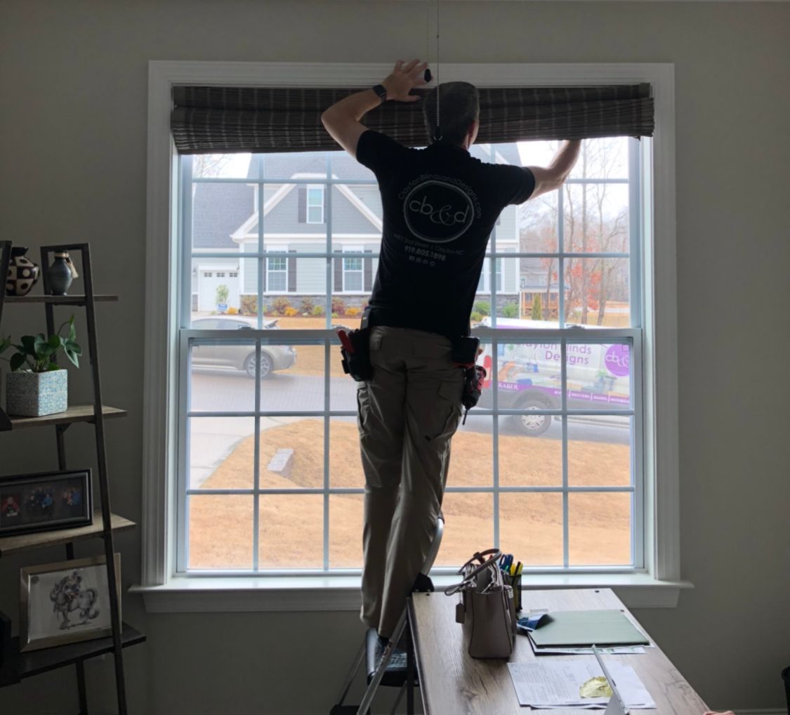 Person installing window blinds, standing on a step ladder inside a room with a large window.