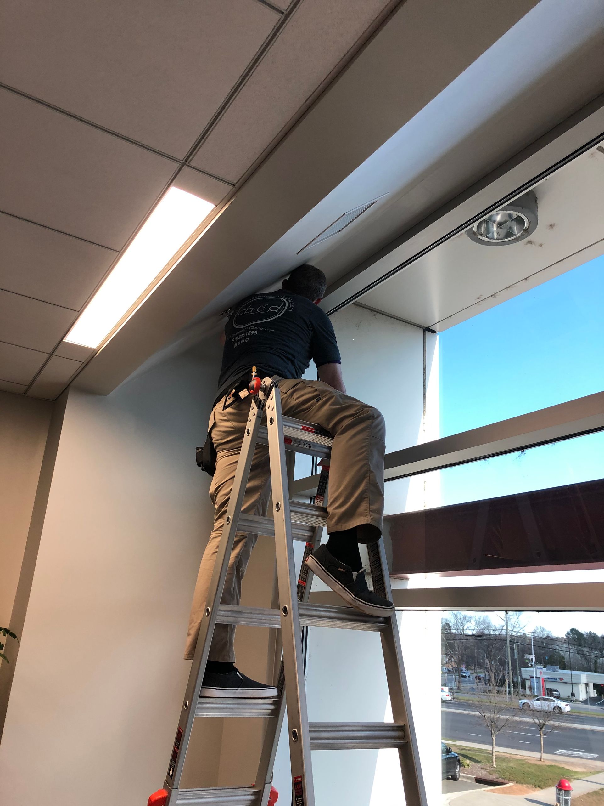 Person on a ladder, installing something above a window. Bright sky outside.