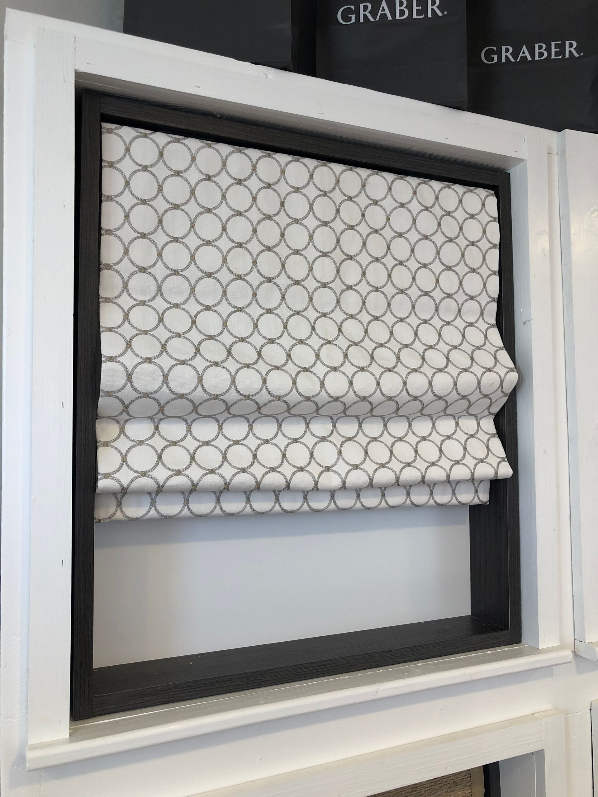 Roman shade with a white background and grey circle pattern, framed in a white window with a dark grey interior.