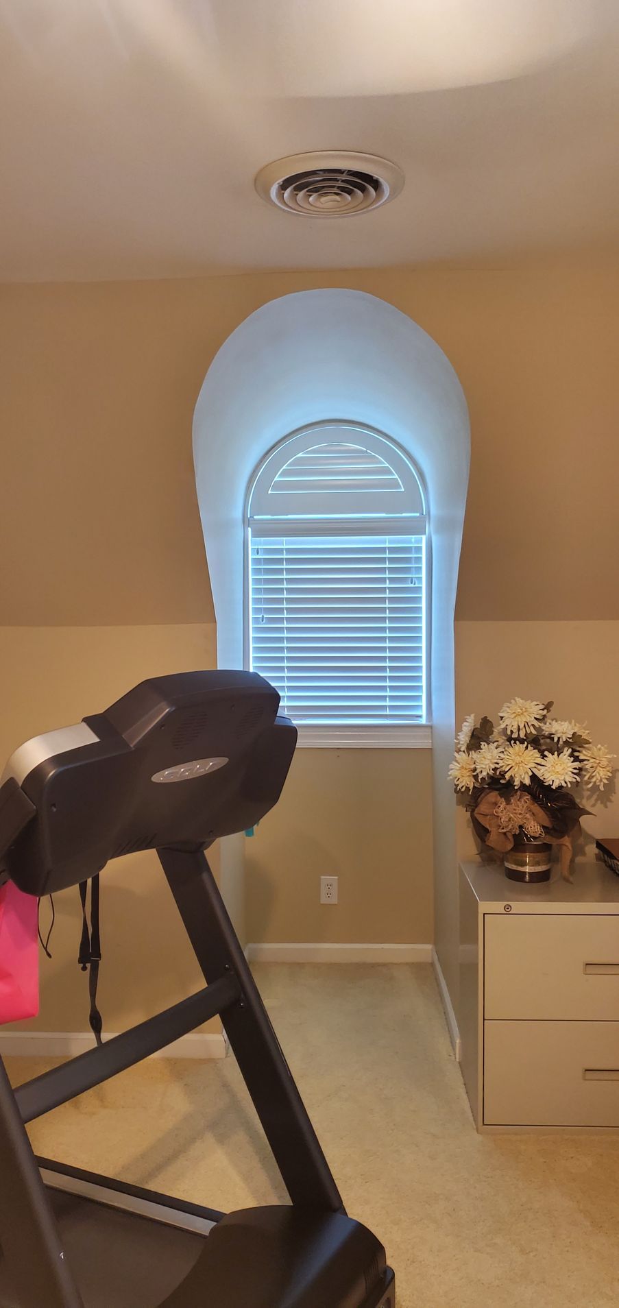 Treadmill in a room with a window, white blinds, and floral arrangement on a dresser.