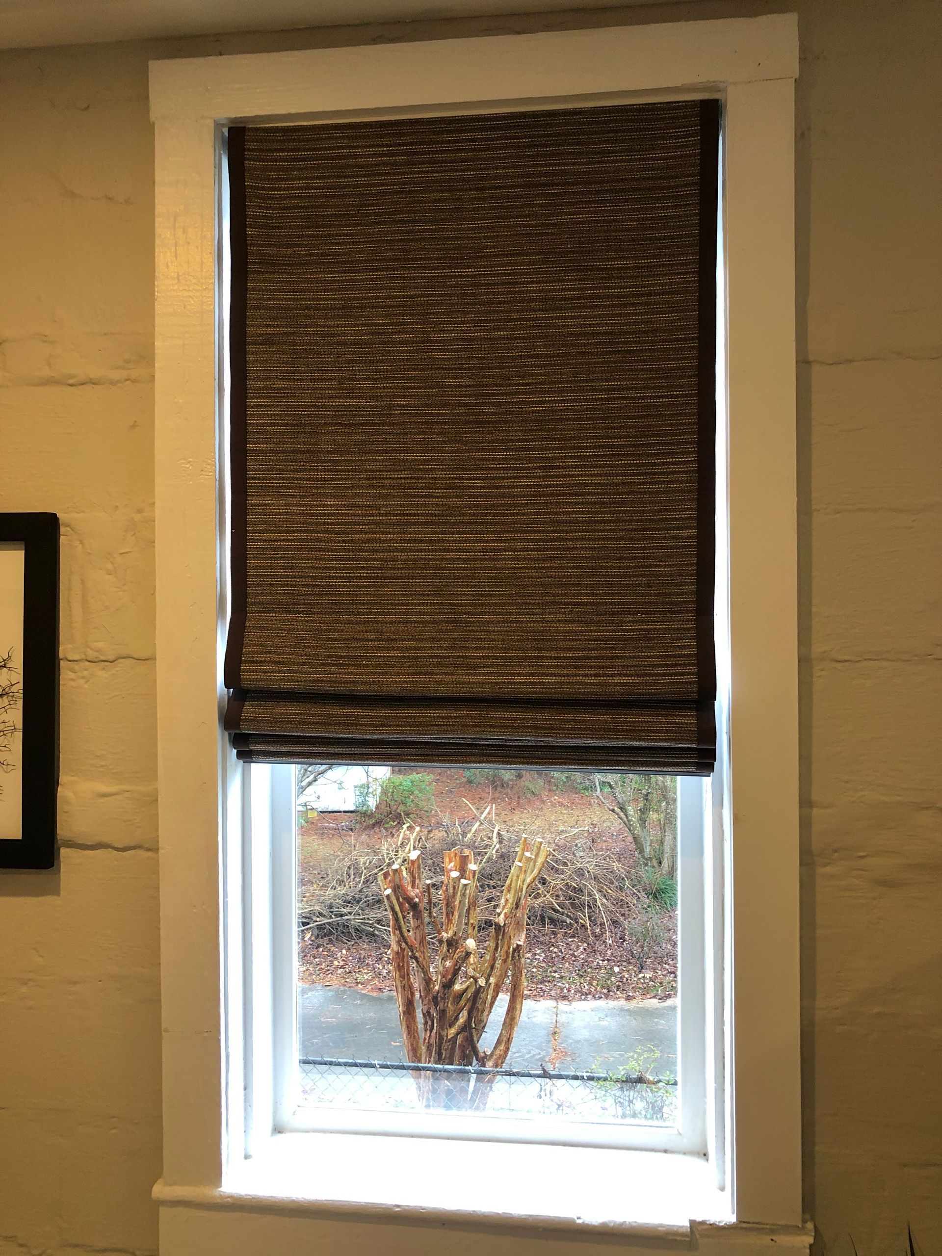 Window with a brown woven shade, white frame, and a view of greenery outside.