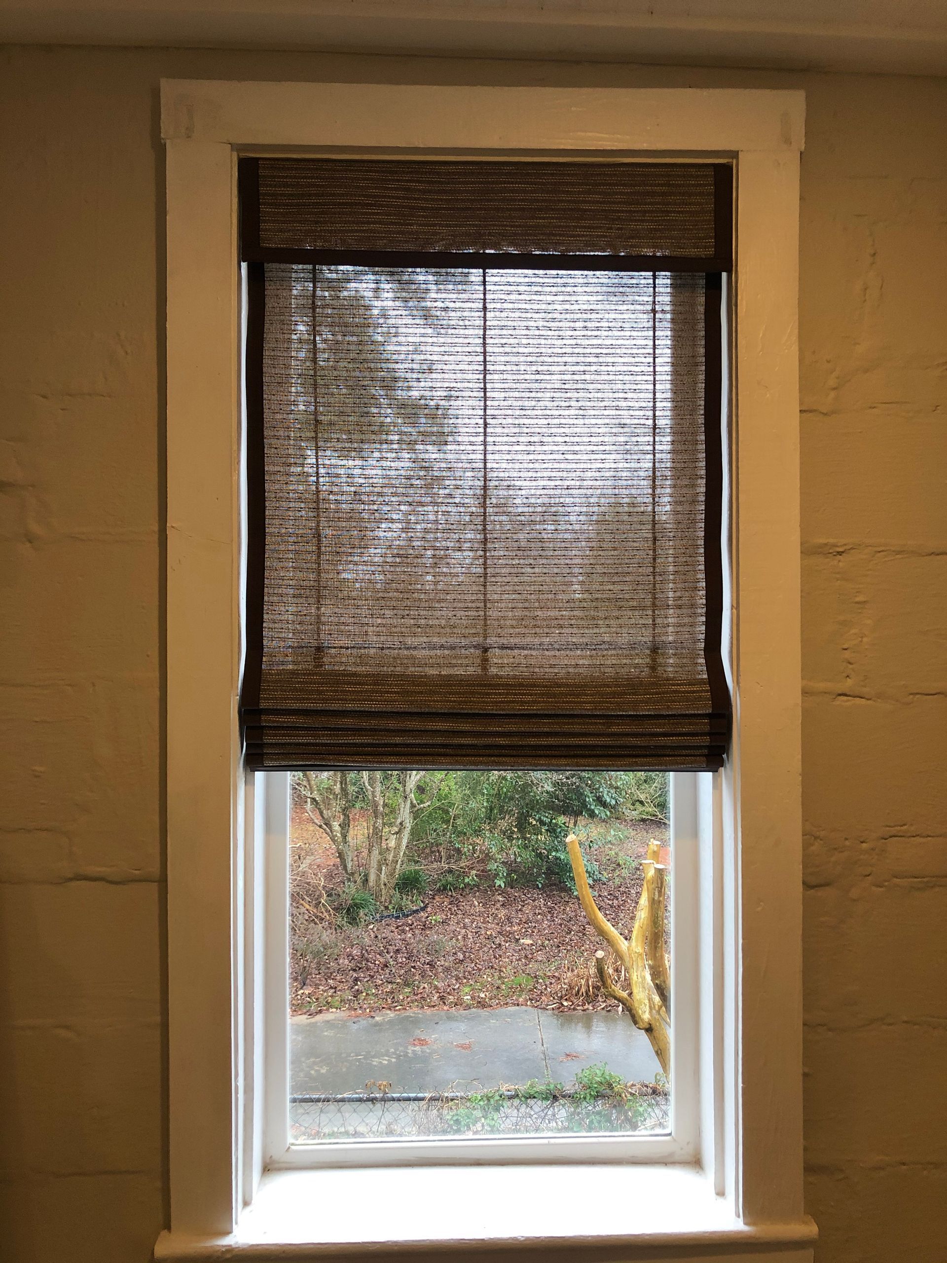 Window with woven shade, view of a yard with autumn leaves, white trim, beige wall.