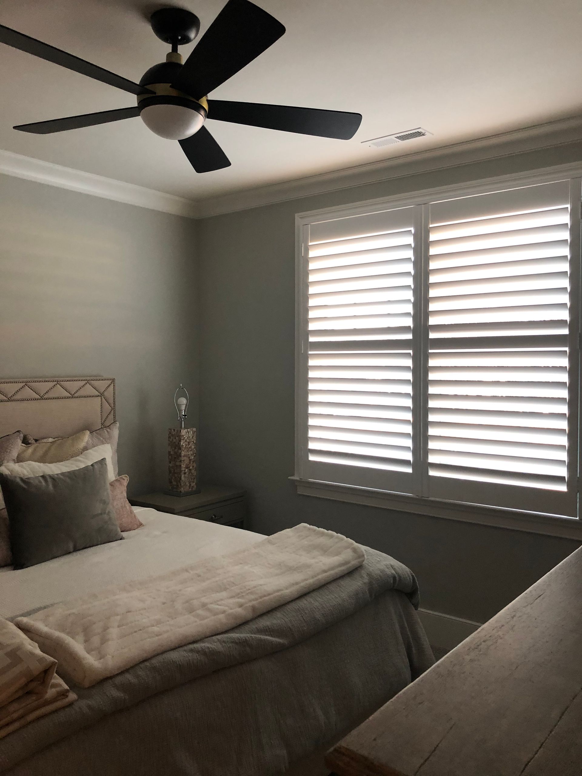 Bedroom with gray walls, white shutters, bed with pillows, and a ceiling fan.