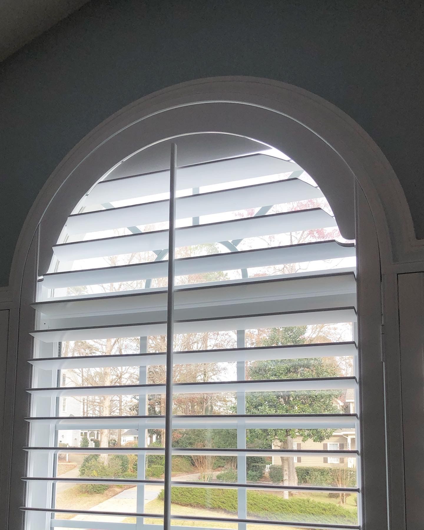 White plantation shutters on arched window. Trees and houses visible through slats.