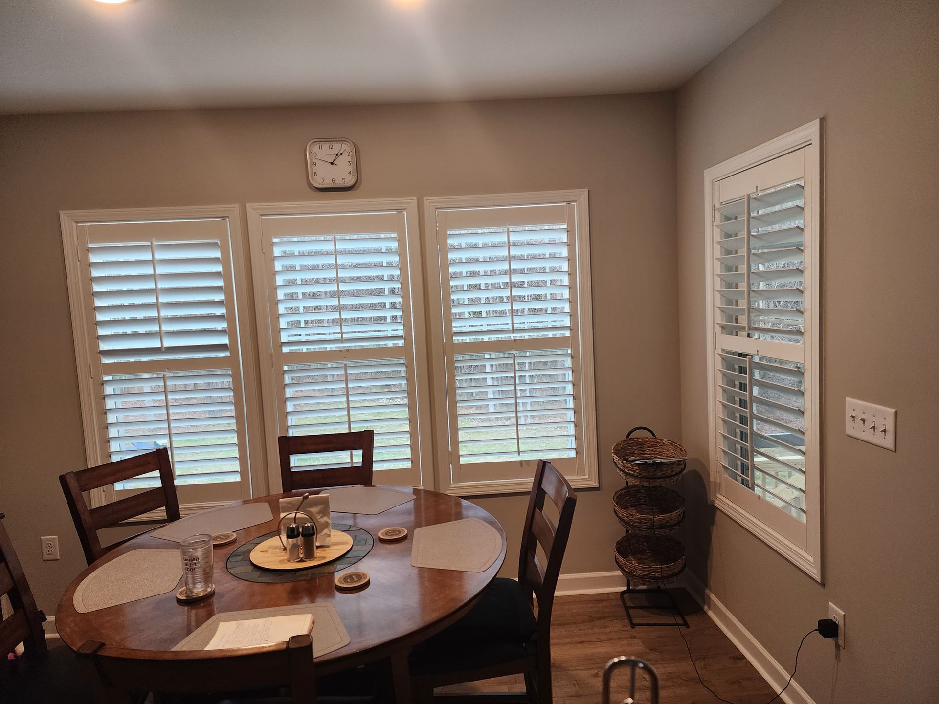 A dining room with a round wooden table, three windows with shutters, and a wall clock.