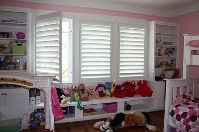 Pink bedroom with white shutters, built-in shelves, and window seat holding stuffed animals.