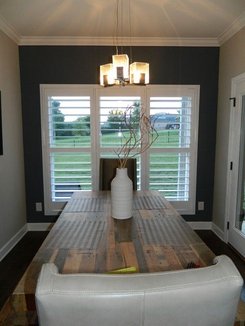 Dining room with wooden table, white shutters, and hanging light fixture. Gray accent wall and white chair.