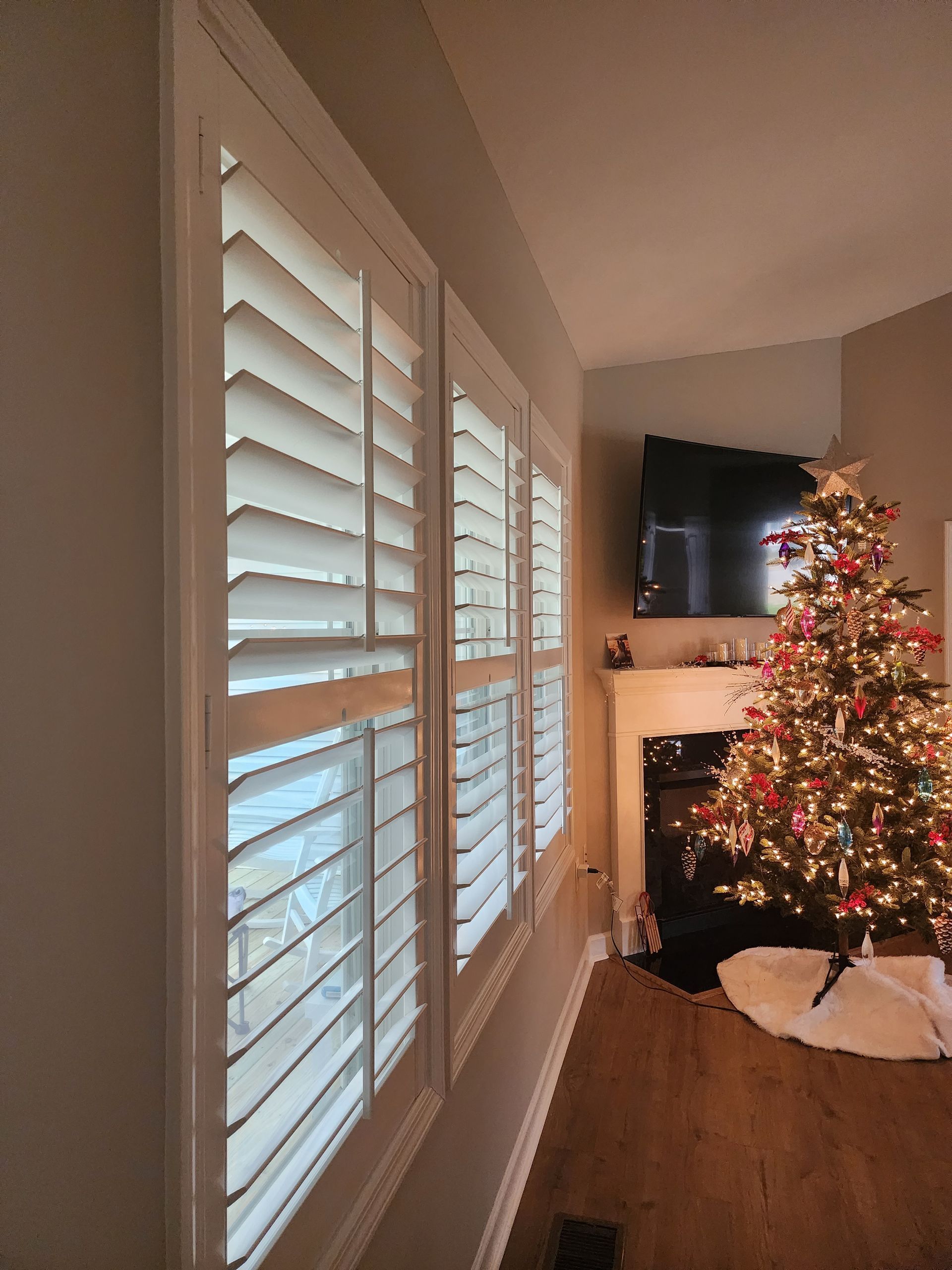 White shutters on windows, Christmas tree decorated with lights next to fireplace.