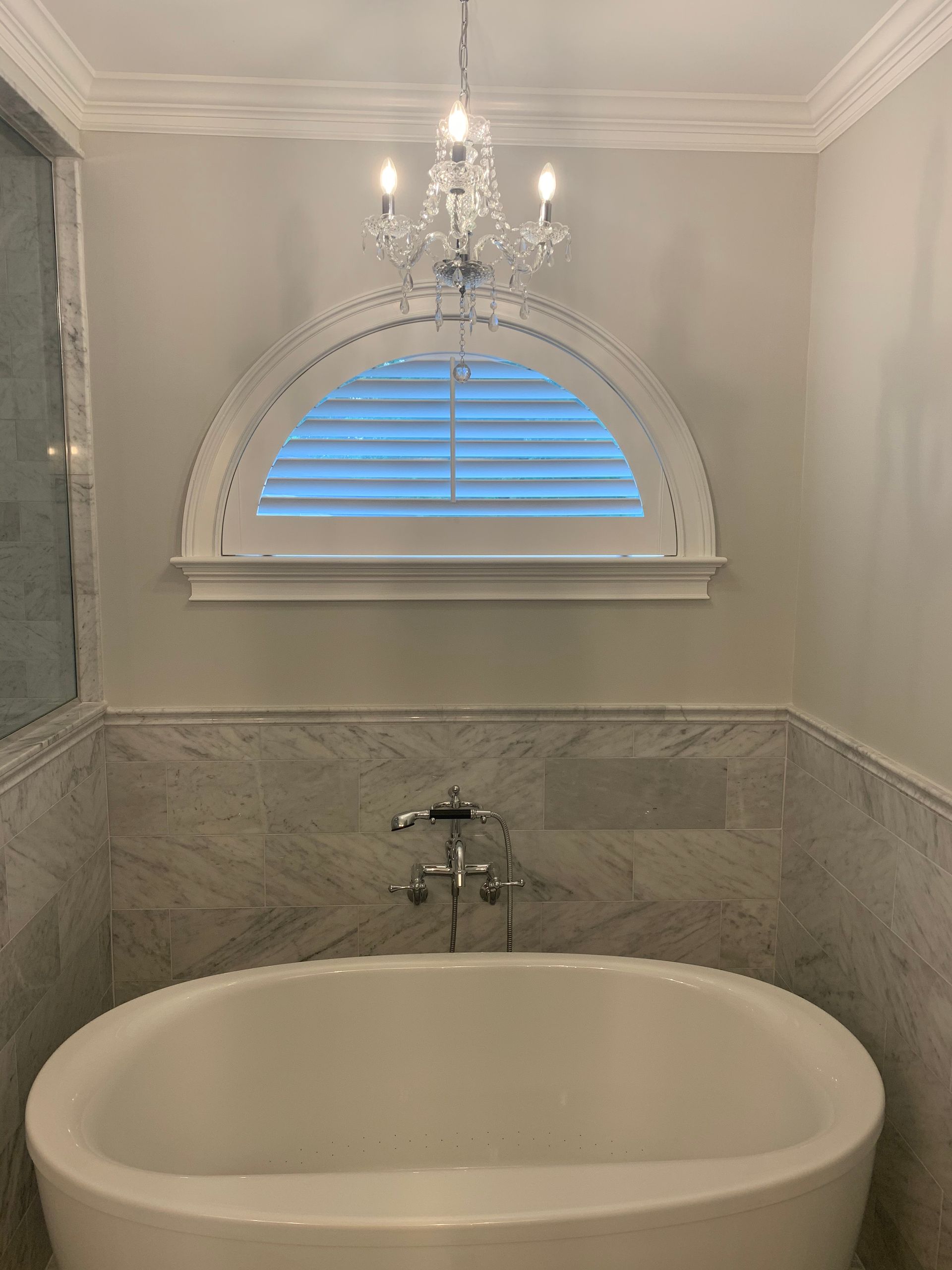 Bathroom with white oval tub, marble tile, arched window with shutters, and crystal chandelier.