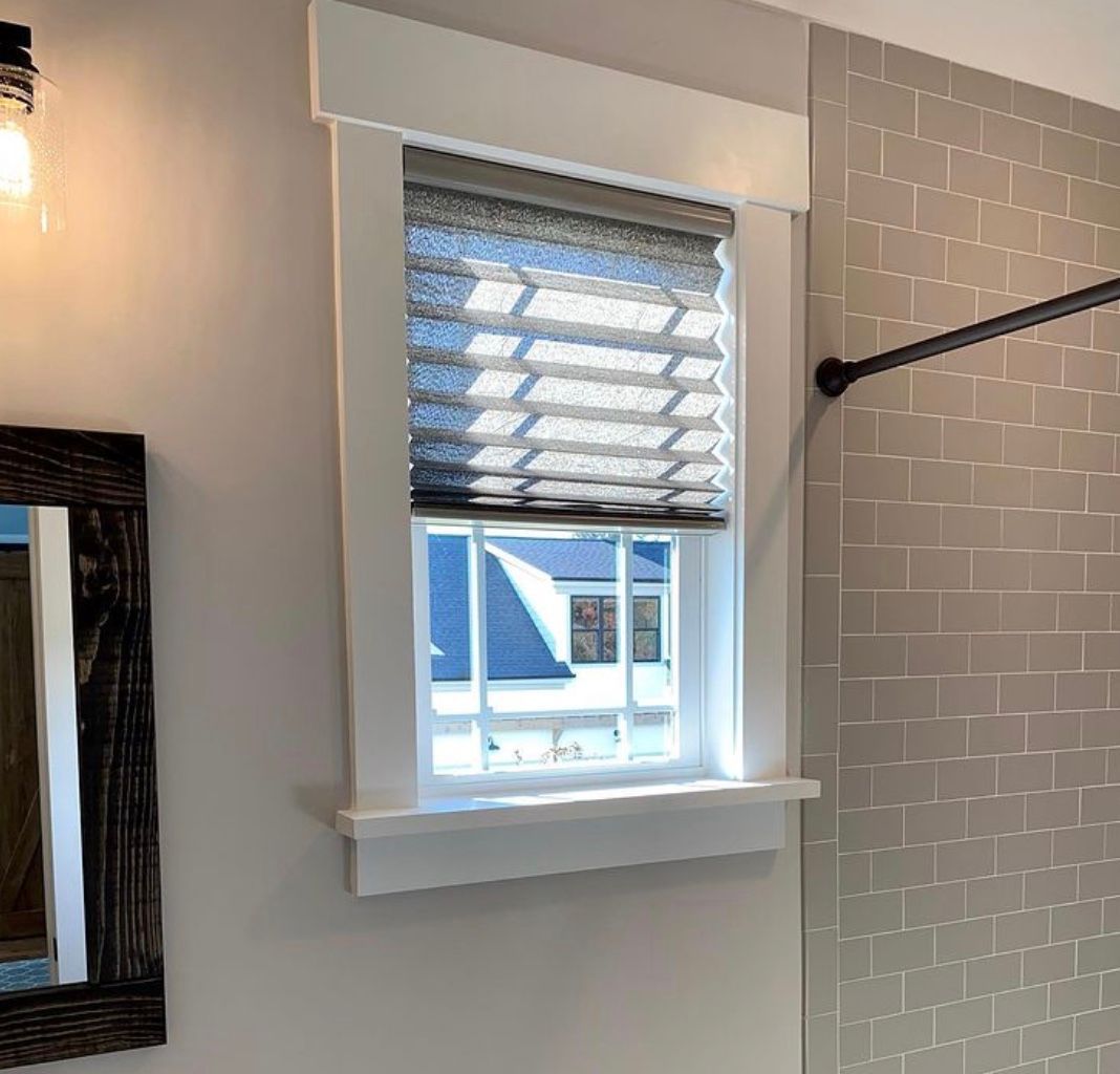 Window with gray pleated shade, white trim, in a bathroom with gray tile and a black shower rod.