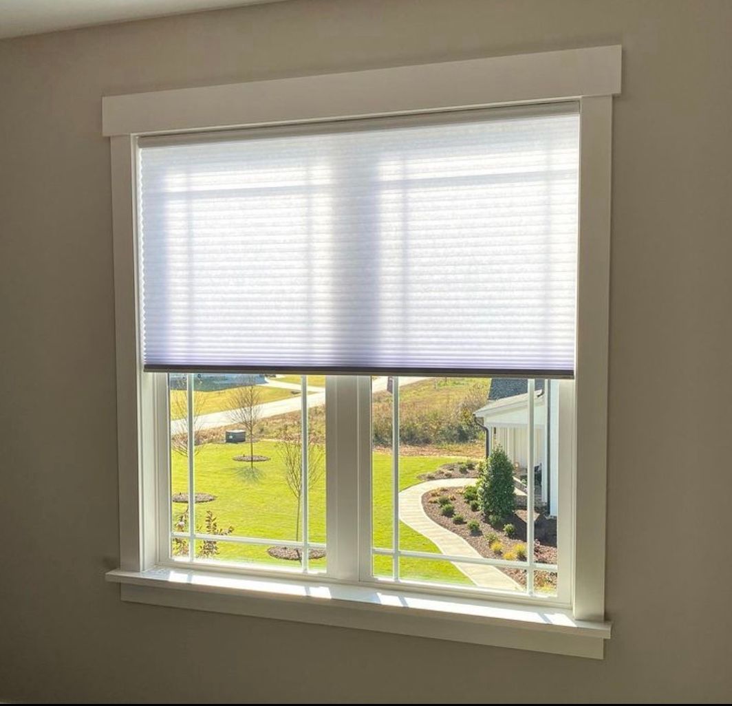 Window with white trim and a white pleated shade, overlooking a grassy yard and pathway.