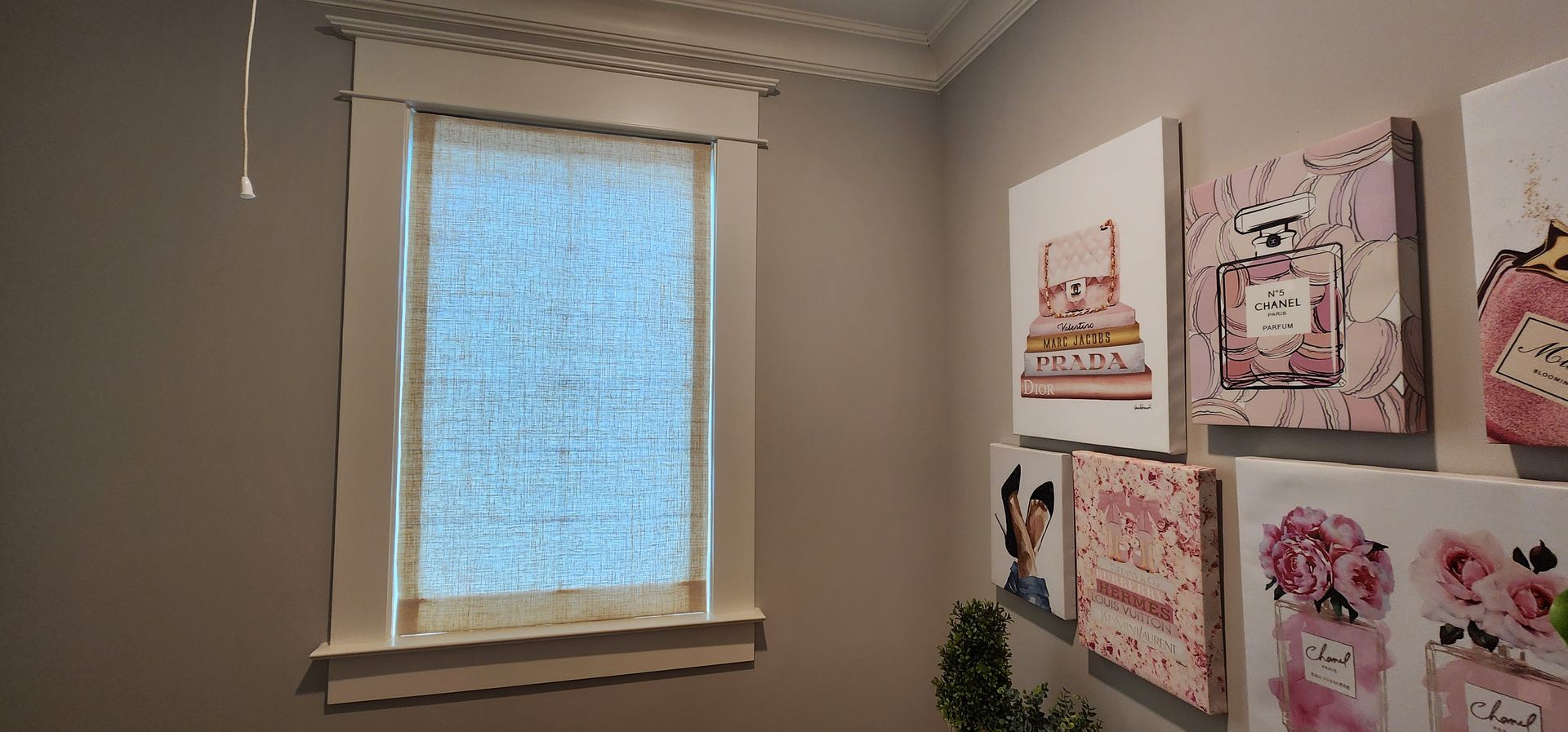 Room with a window, white trim, and light blue window covering. Wall art with pink perfume bottles.