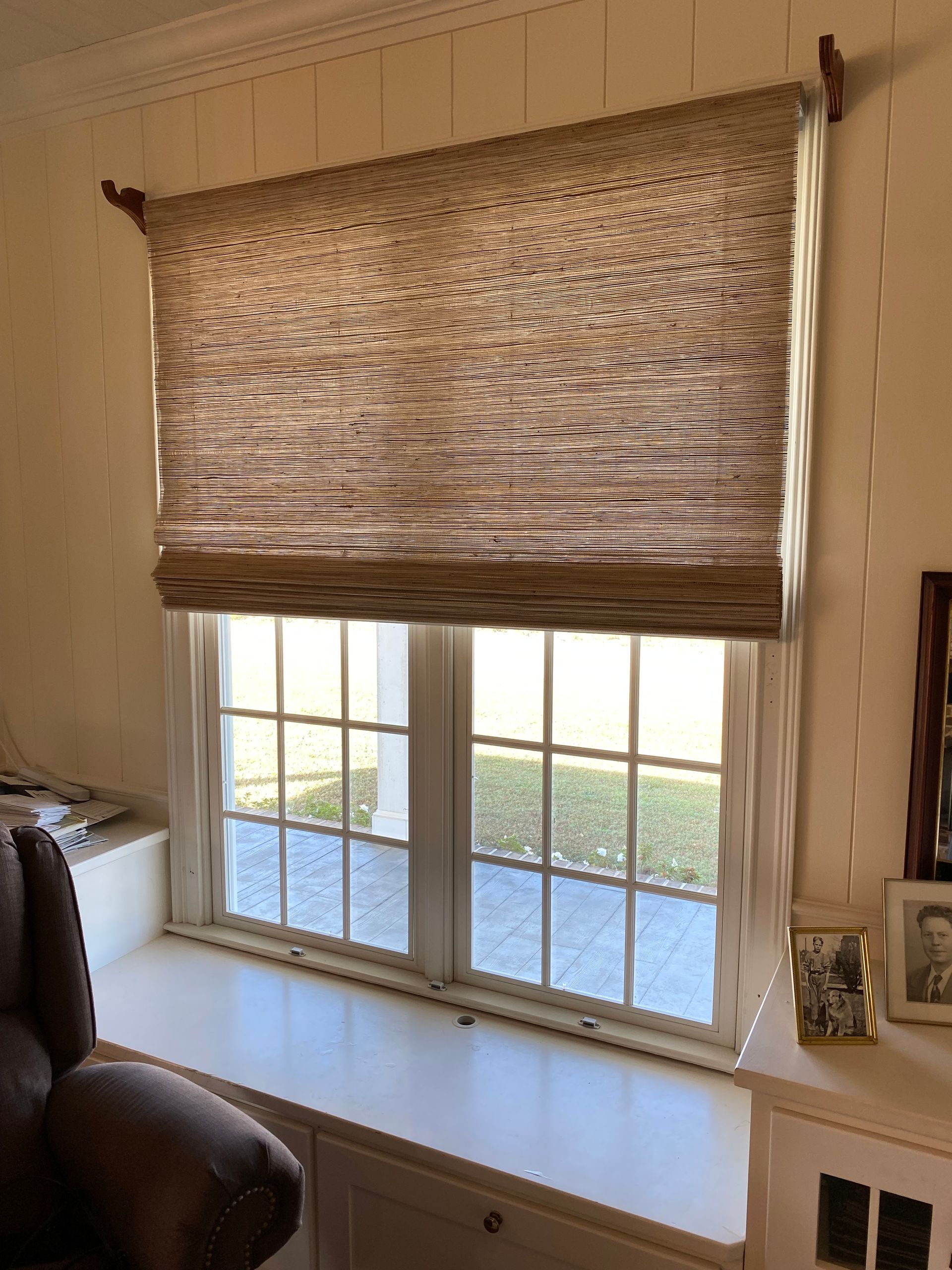 Roman shade covering window; tan fabric, wooden rod. White walls, window seat.