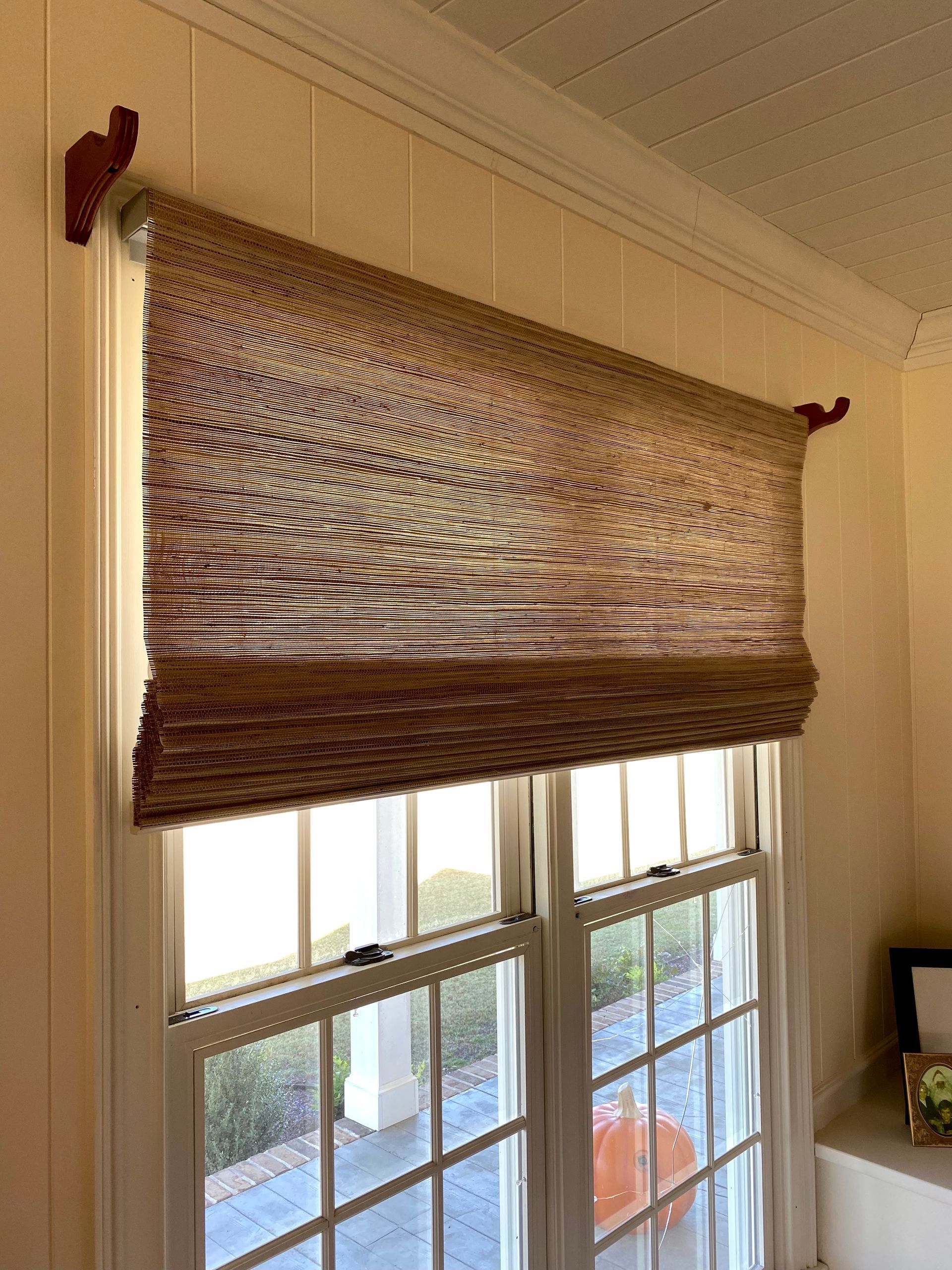 Woven window shade, brown and beige, above a white-framed window. Wooden decorative brackets.