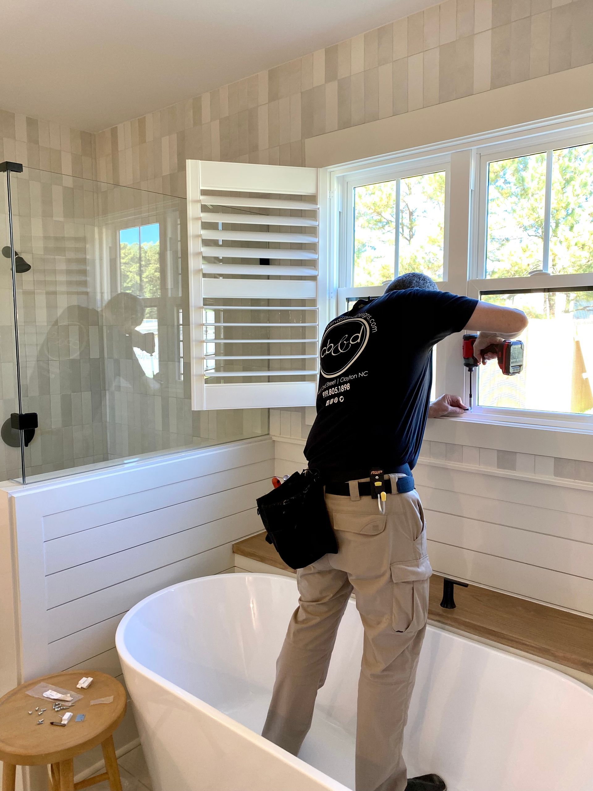 Person installing window shutters in a bathroom with a white tub and wood accents.