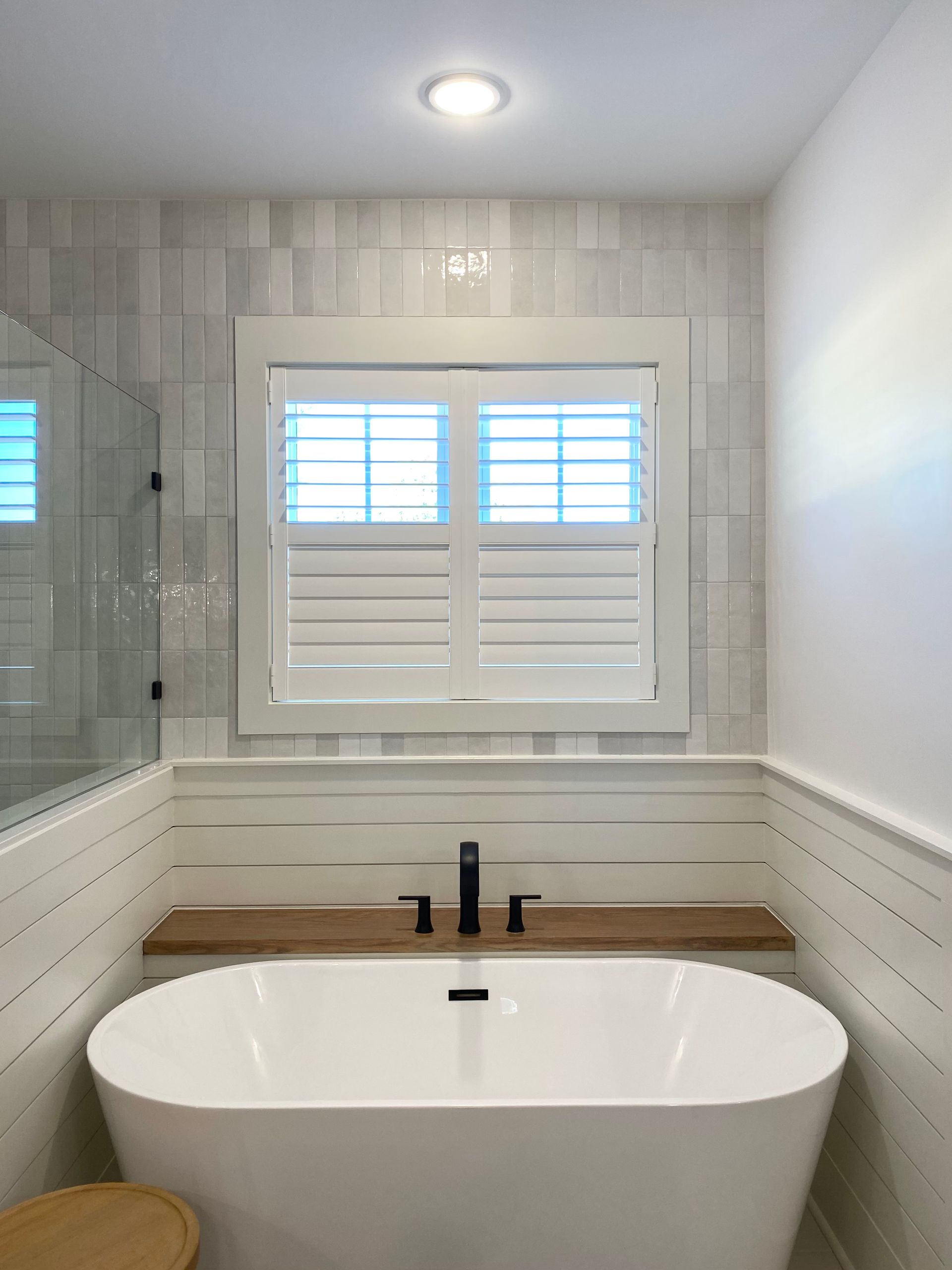 Bathroom with a white freestanding tub, wood shelf, black faucet, white shutters, and tiled walls.