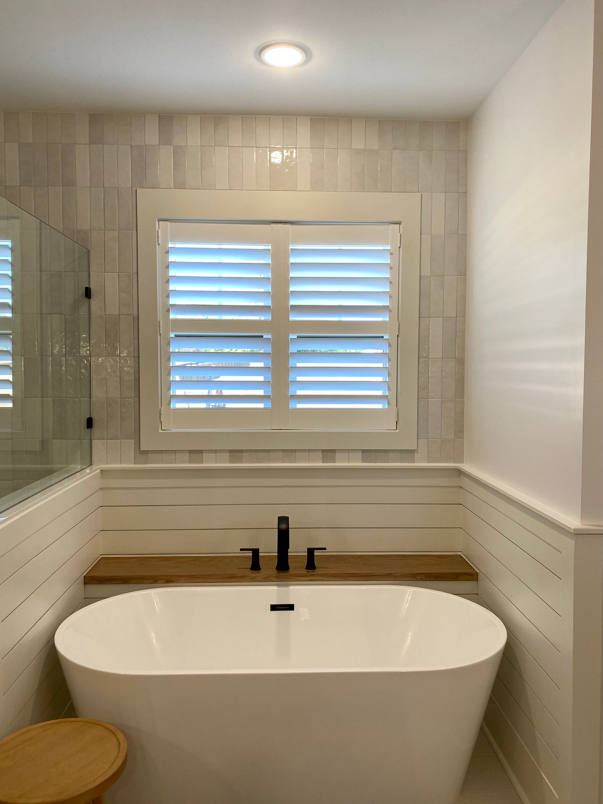 Freestanding white bathtub below a window with white shutters in a bathroom.