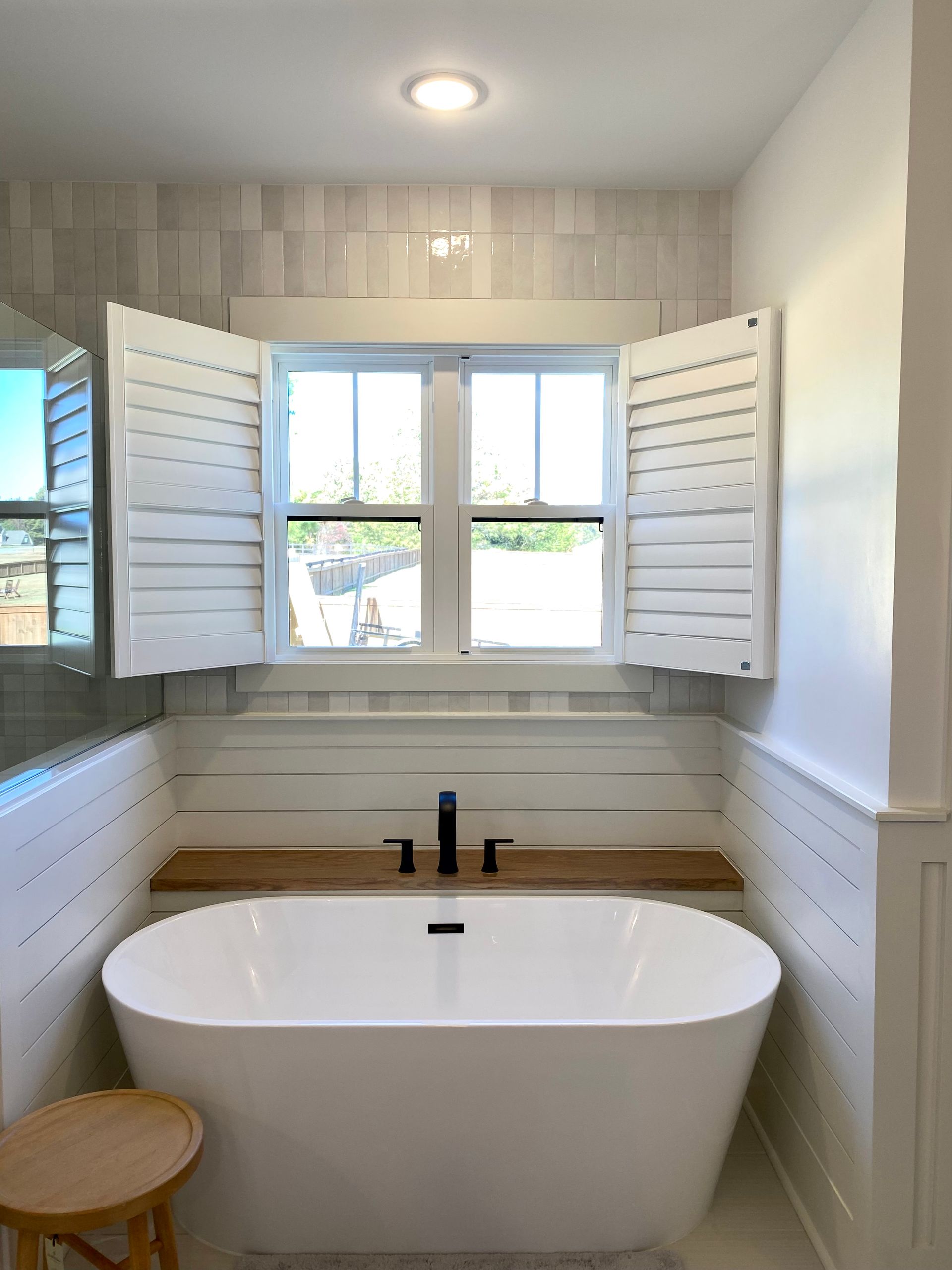 Freestanding white bathtub under a window with open shutters. Black faucet, wooden shelf, and a small stool.