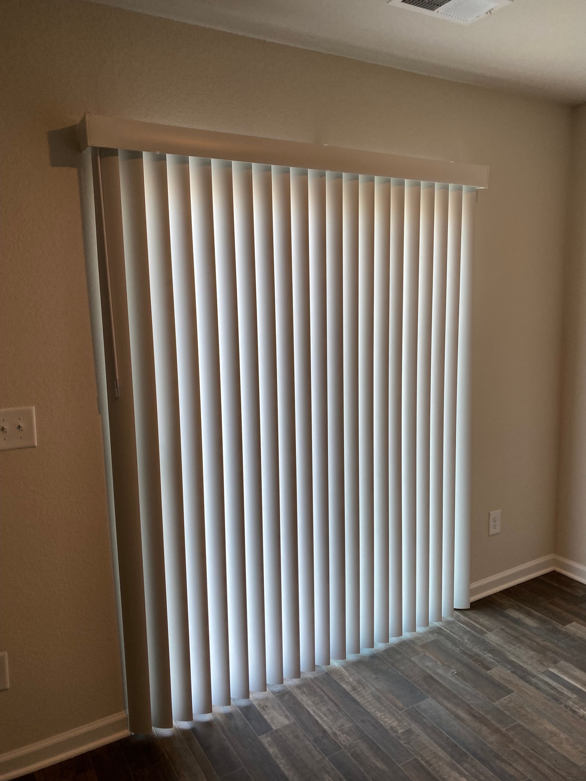 Vertical blinds covering a sliding glass door in a room with wood-look flooring and cream-colored walls.
