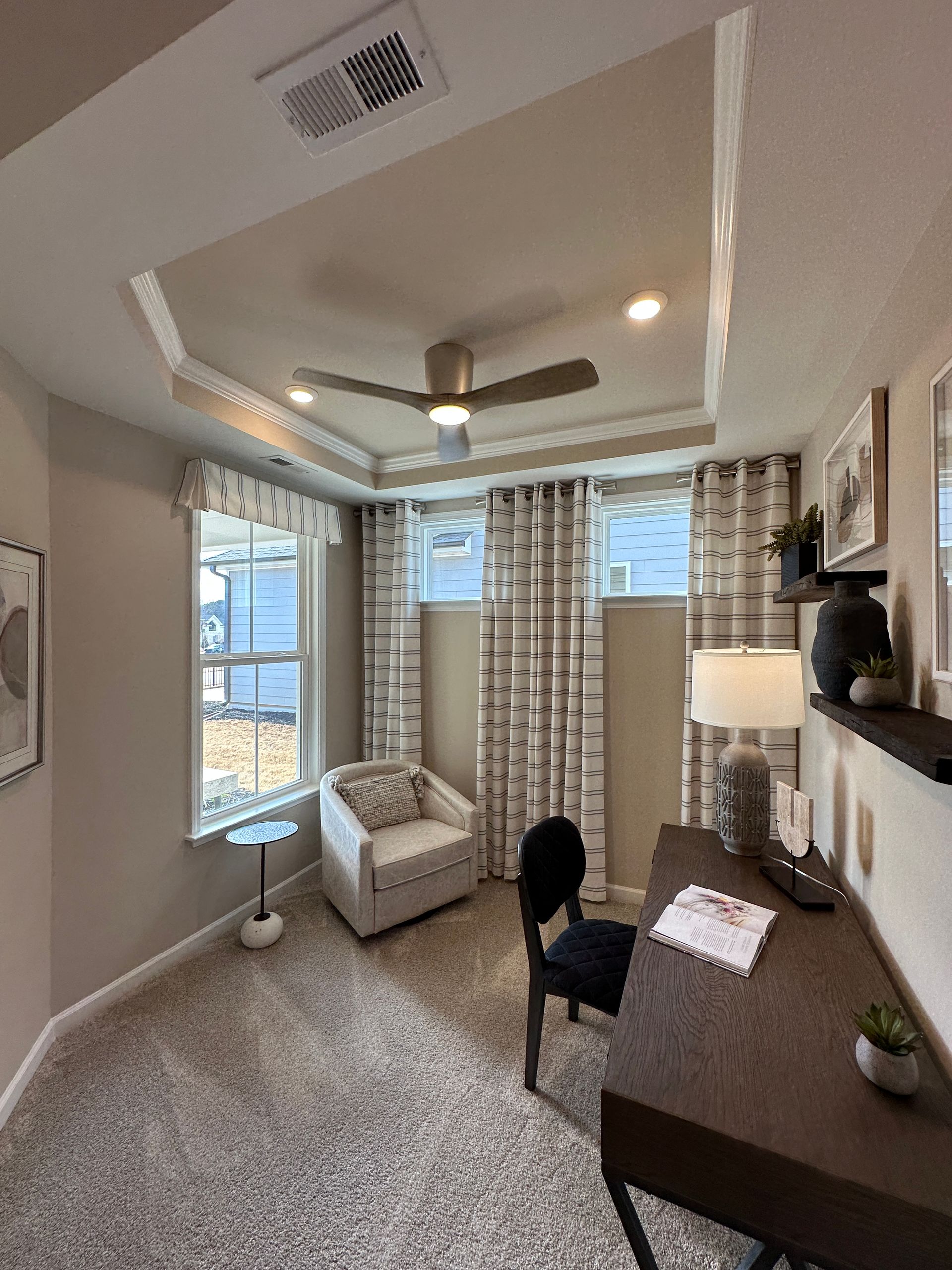 Home office with desk, chair, armchair, and patterned curtains. Beige walls, ceiling fan, and carpet.