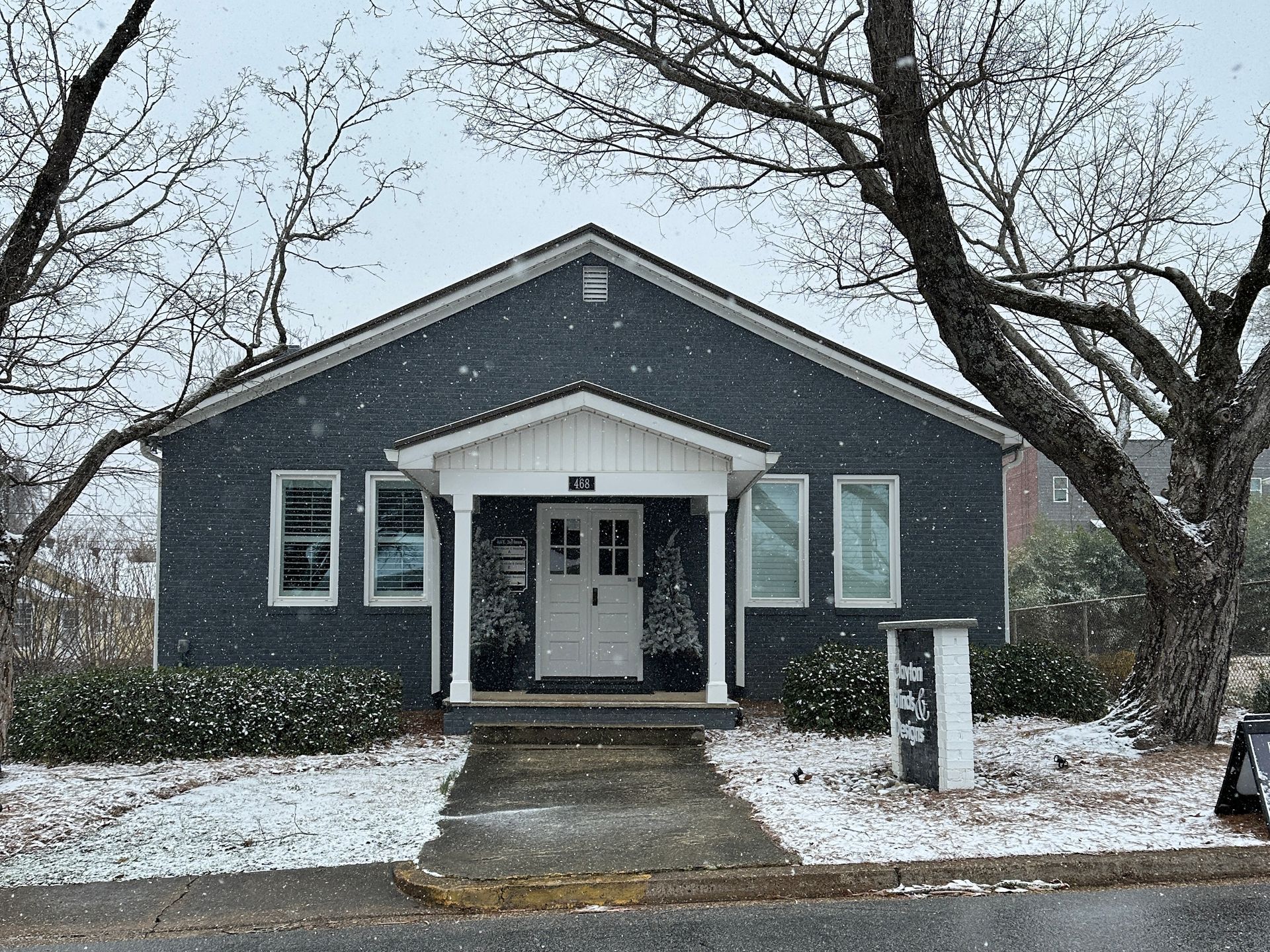 Snowy exterior view of a dark blue building with a white door and porch, framed by bare trees.