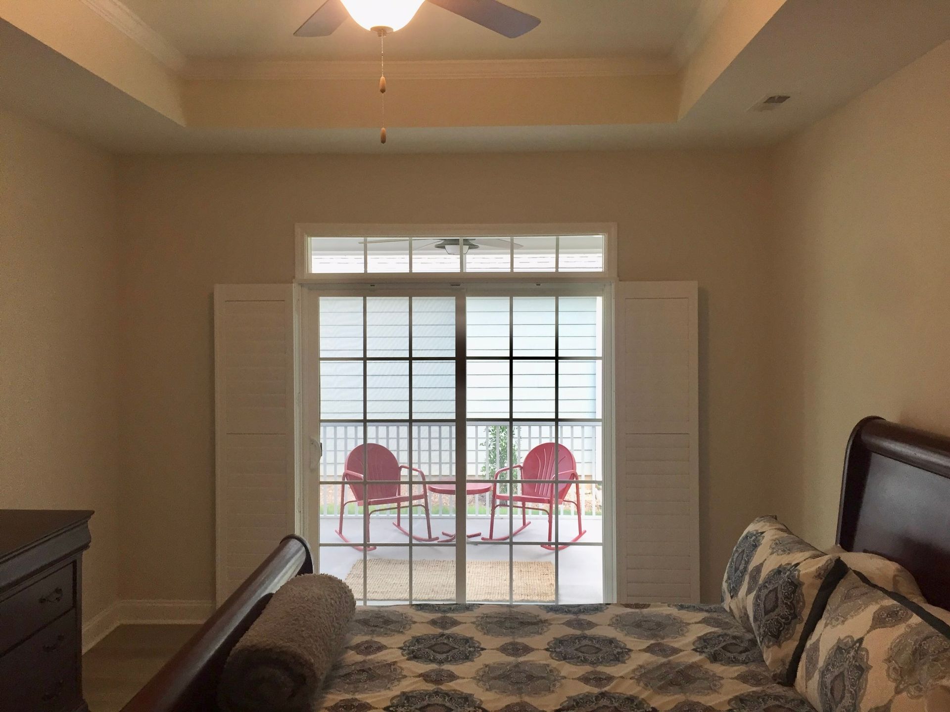 Bedroom with large sliding glass door opening to a balcony with red chairs. White shutters frame the door.