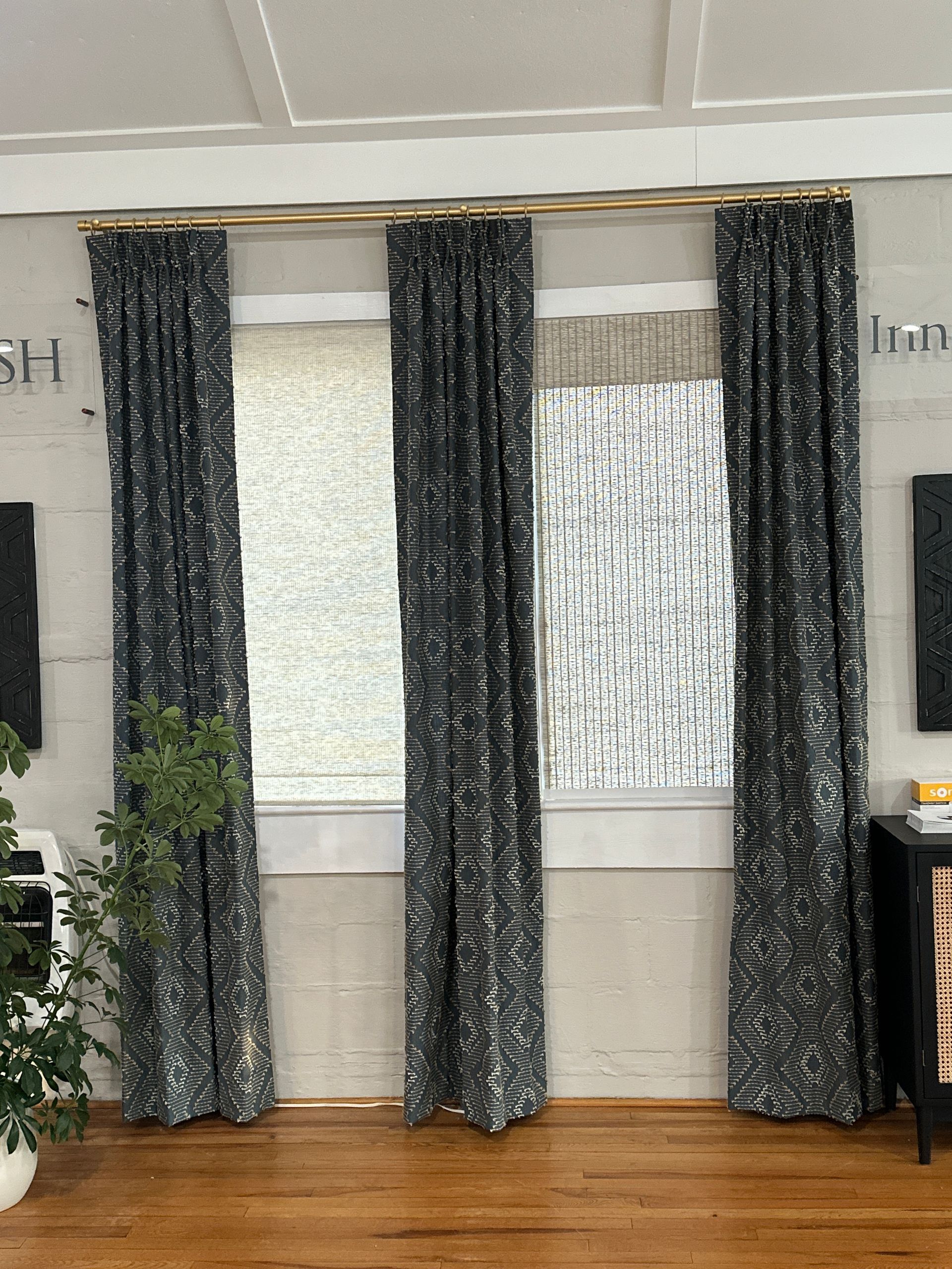 Curtains framing three windows with textured light-colored shades. Wooden floors, a plant, and a cabinet are in the room.