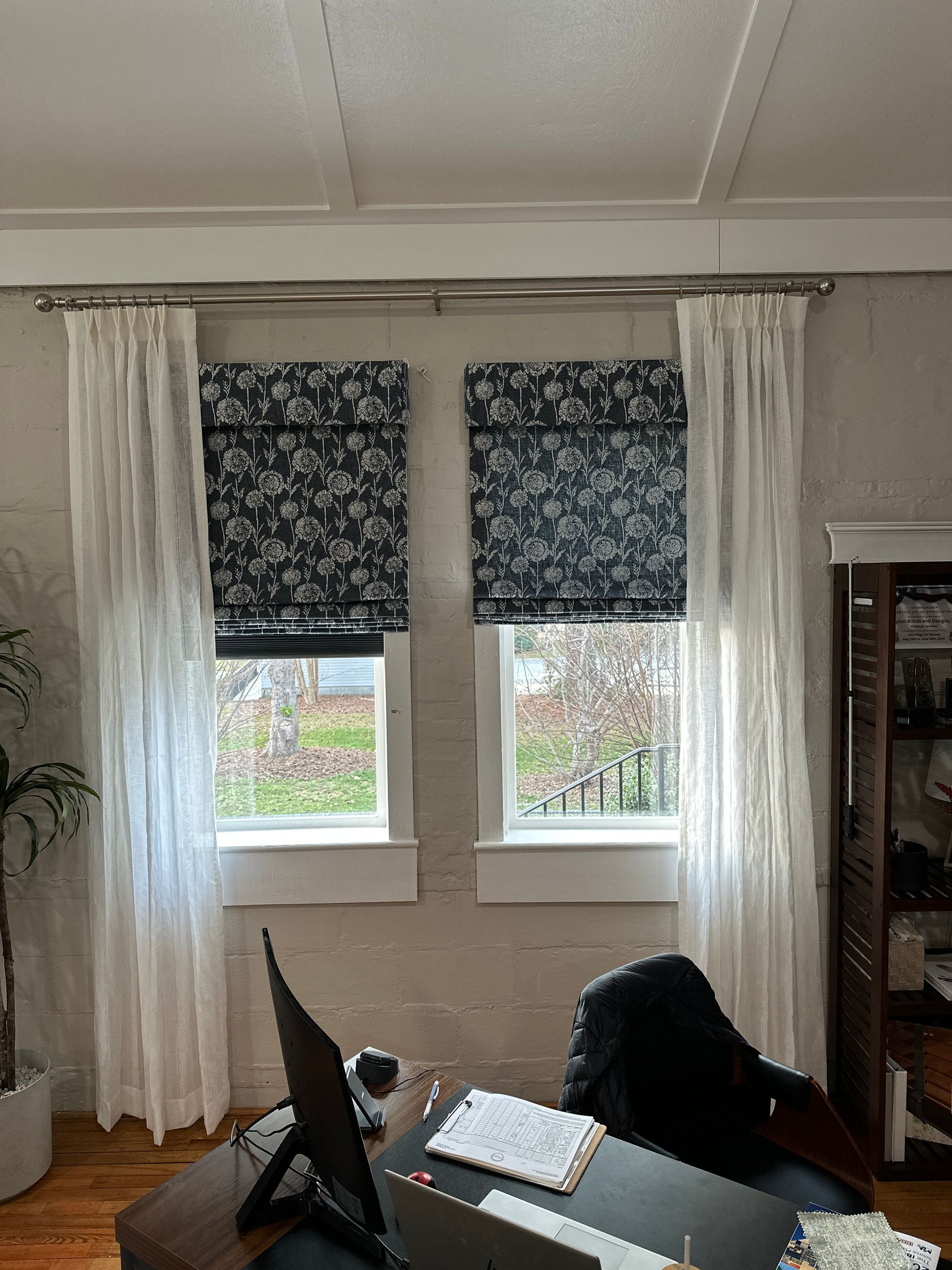 White curtains flank windows with black floral Roman shades; a desk and chair are visible below.