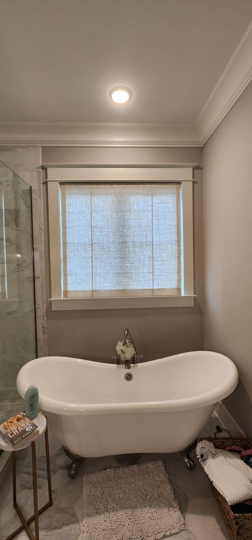 A clawfoot bathtub sits under a window in a bathroom. There is a small table with a book to the left.