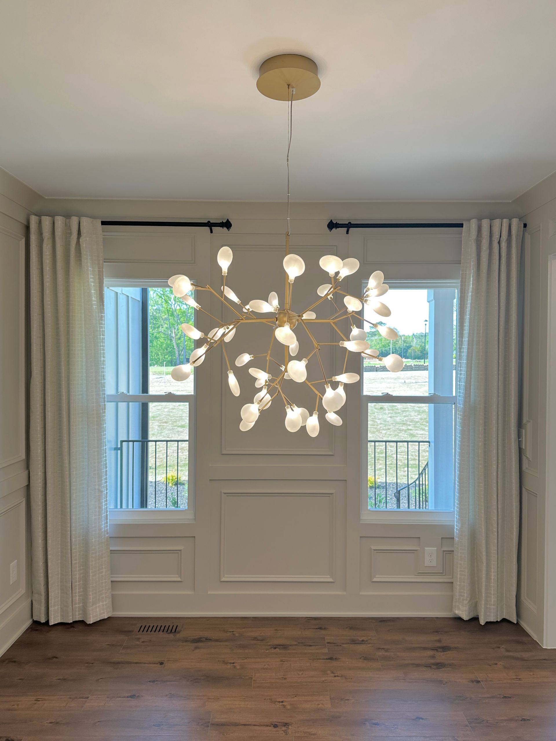 Gold chandelier in a dining room with two windows, sheer white curtains, and wood floors.