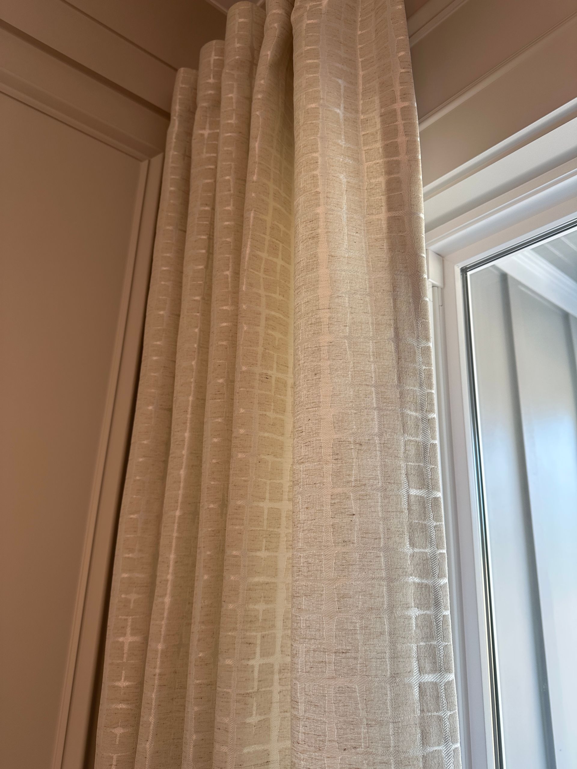 Cream-colored, patterned curtain draped beside a white window frame, showing folded fabric.