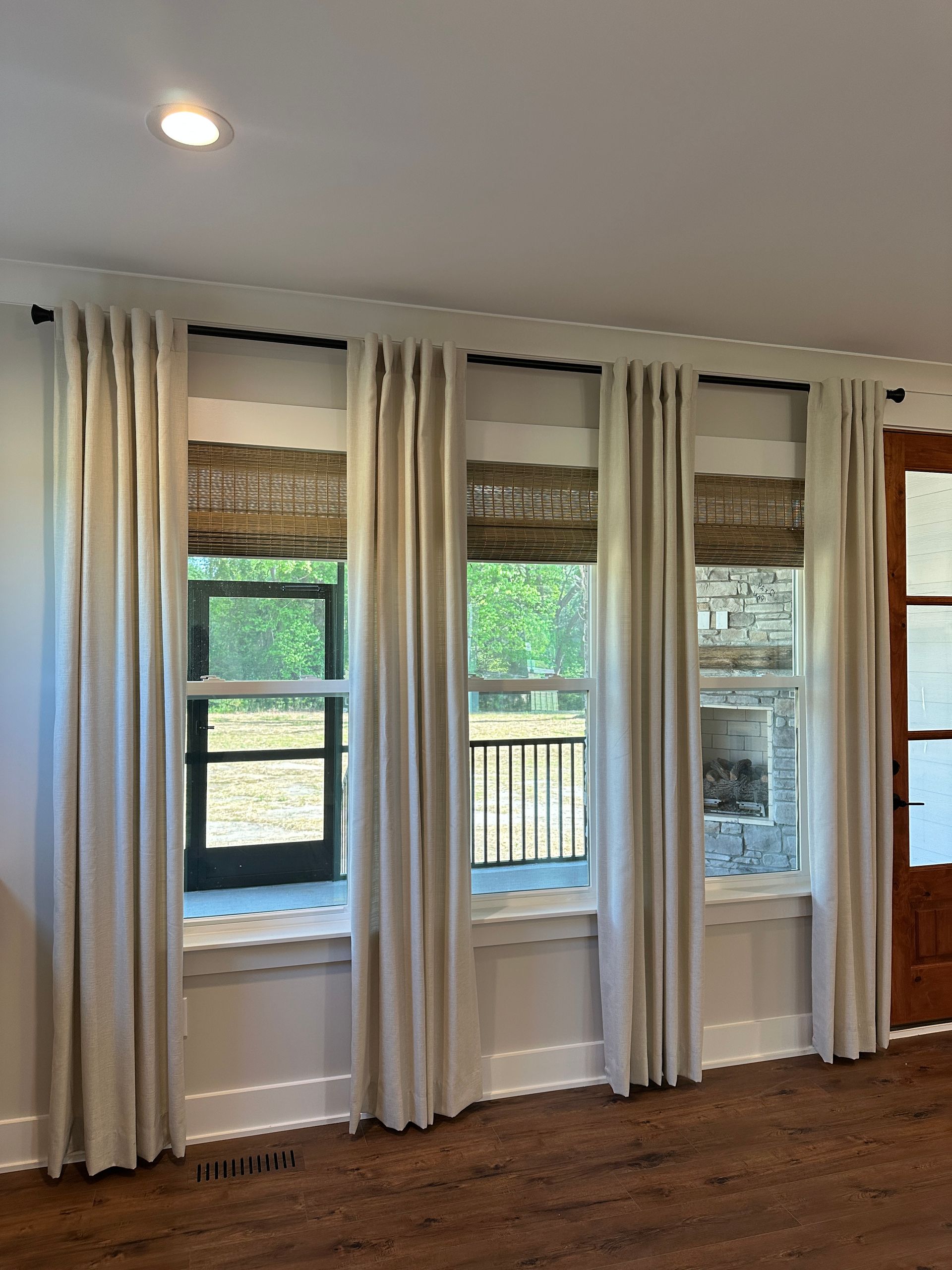 Three windows with beige curtains and woven shades, in a room with hardwood floors.