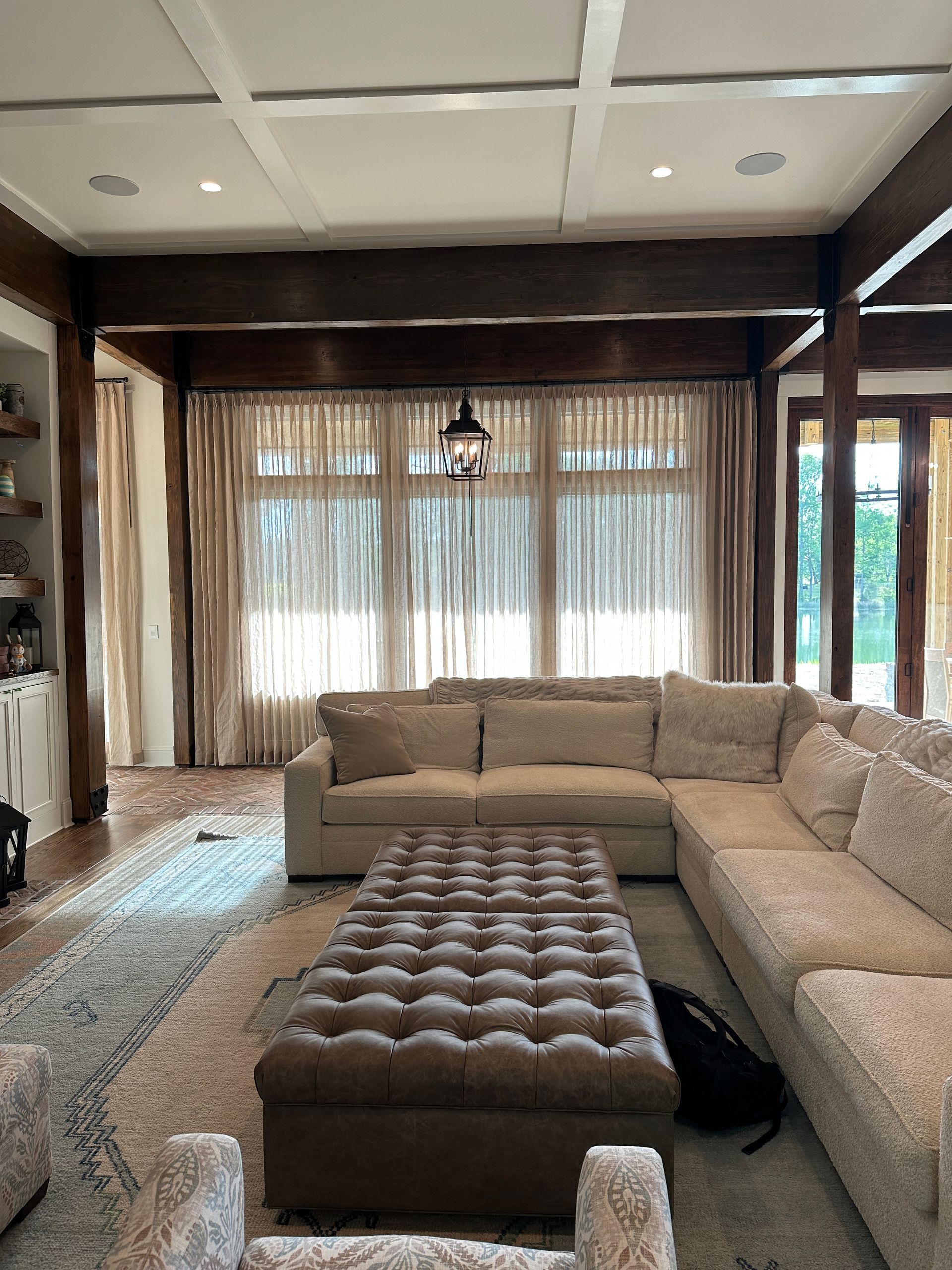 Living room with a beige sectional sofa, a brown ottoman, and sheer curtains. Dark wood beams and a rug.