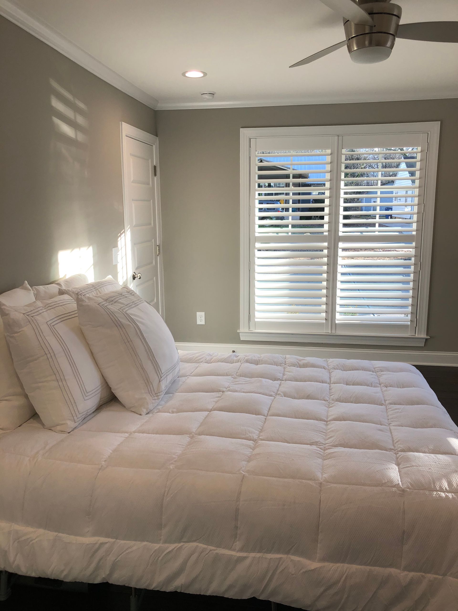Bedroom with white bedding, shutters, and gray walls. Sunlight streams through the window.