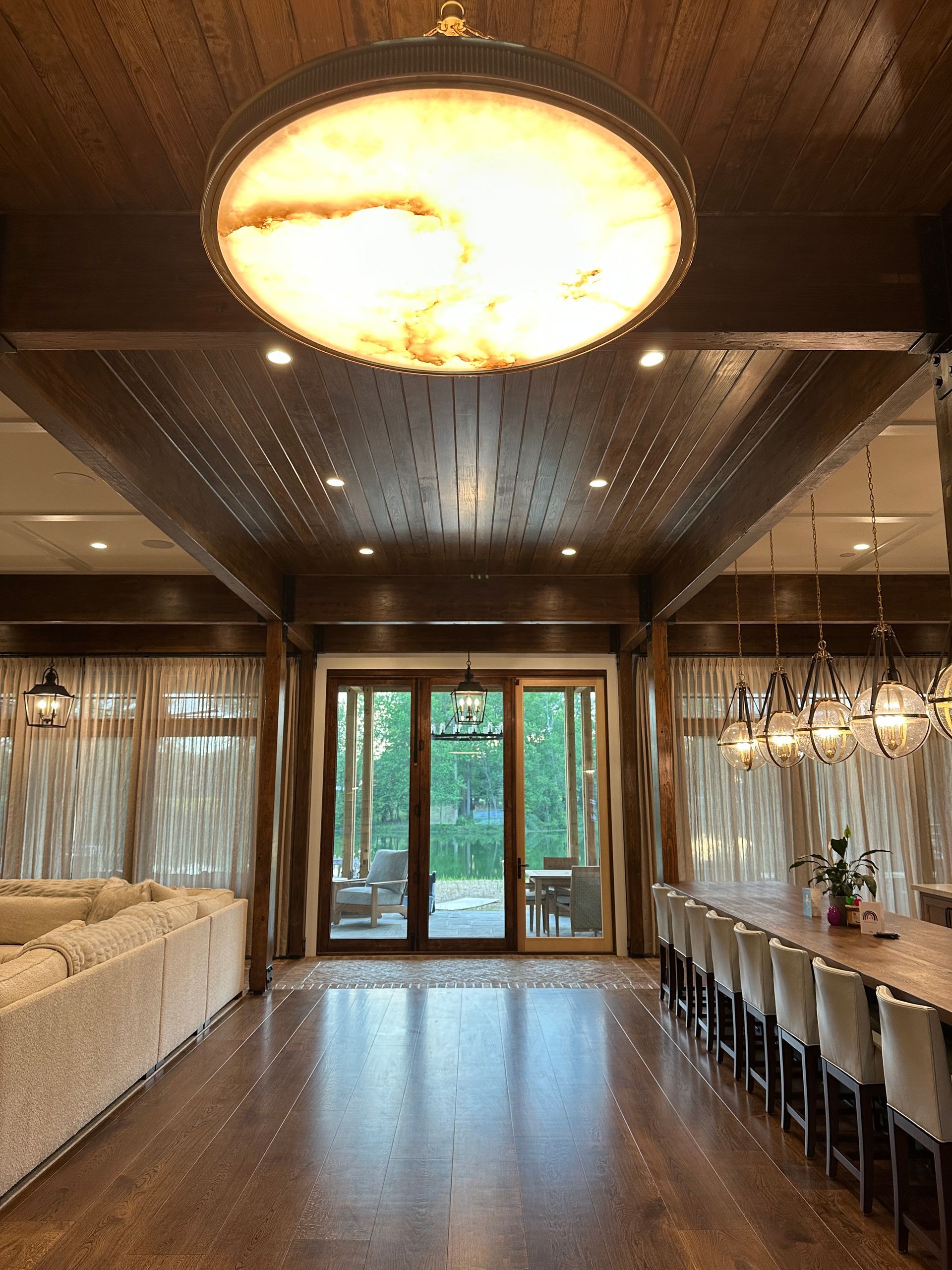 Luxurious dining room with large round light, wooden ceiling, and floor-to-ceiling glass doors.